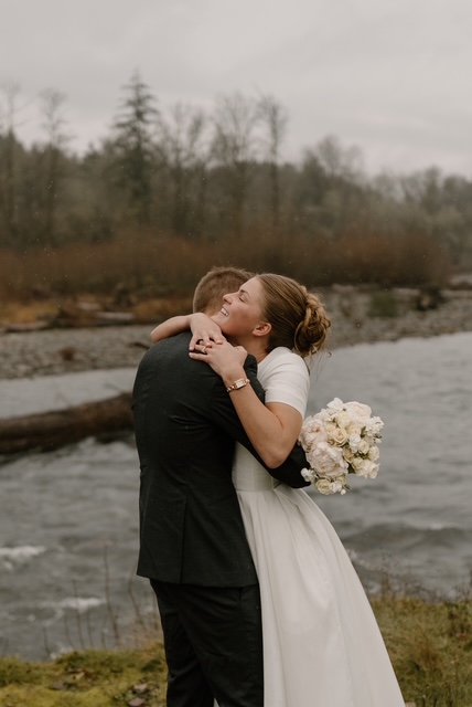 A bride and groom hugging outdoors by a river with trees in the background.