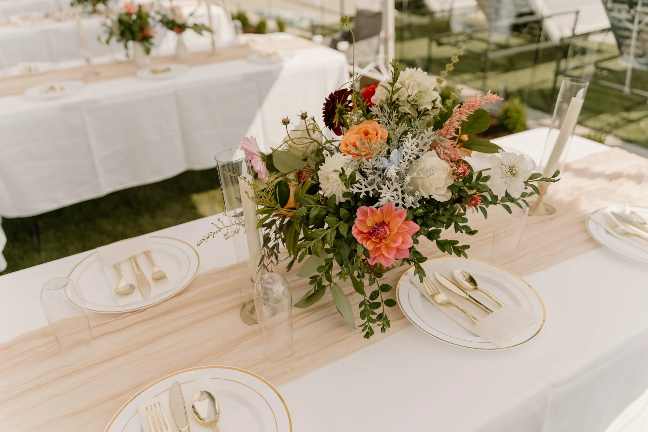 Elegant table setting with a floral centerpiece, gold-rimmed plates, gold cutlery, and glassware on a white tablecloth with a light wooden runner, set outdoors for a special occasion.