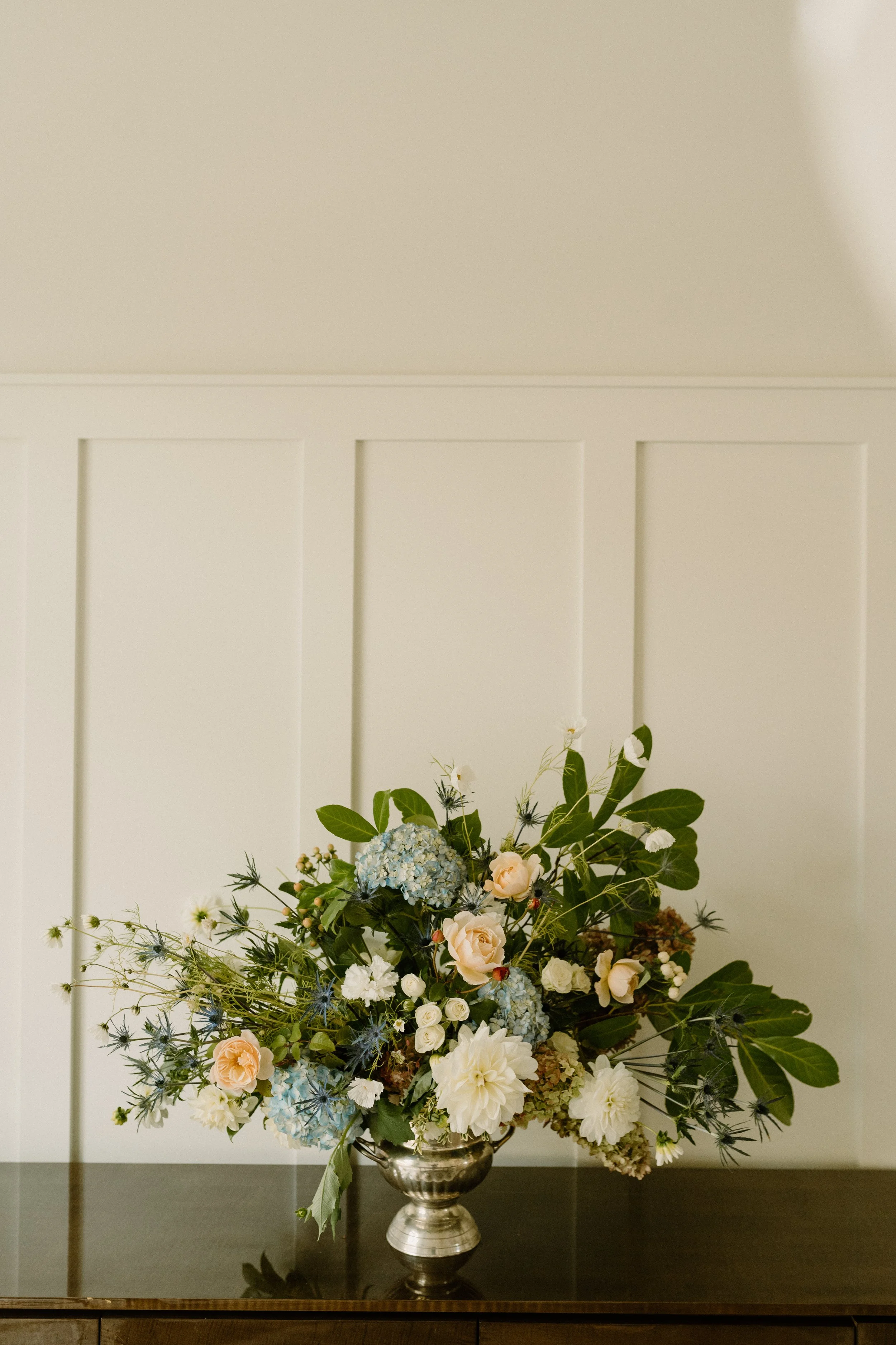 A bouquet of mixed flowers, including blue hydrangeas, white dahlias, peach roses, and various greenery, arranged in a silver vase on a dark wooden surface against a plain white wall.