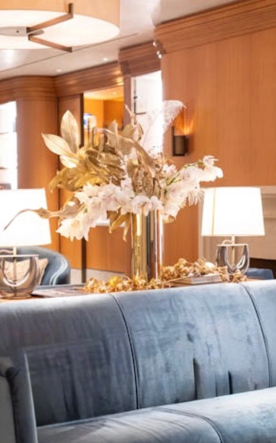 Elegant living room with a large floral arrangement in a tall vase on a table, flanked by lamps and surrounded by leather furniture, wood-paneled walls, and modern decor.