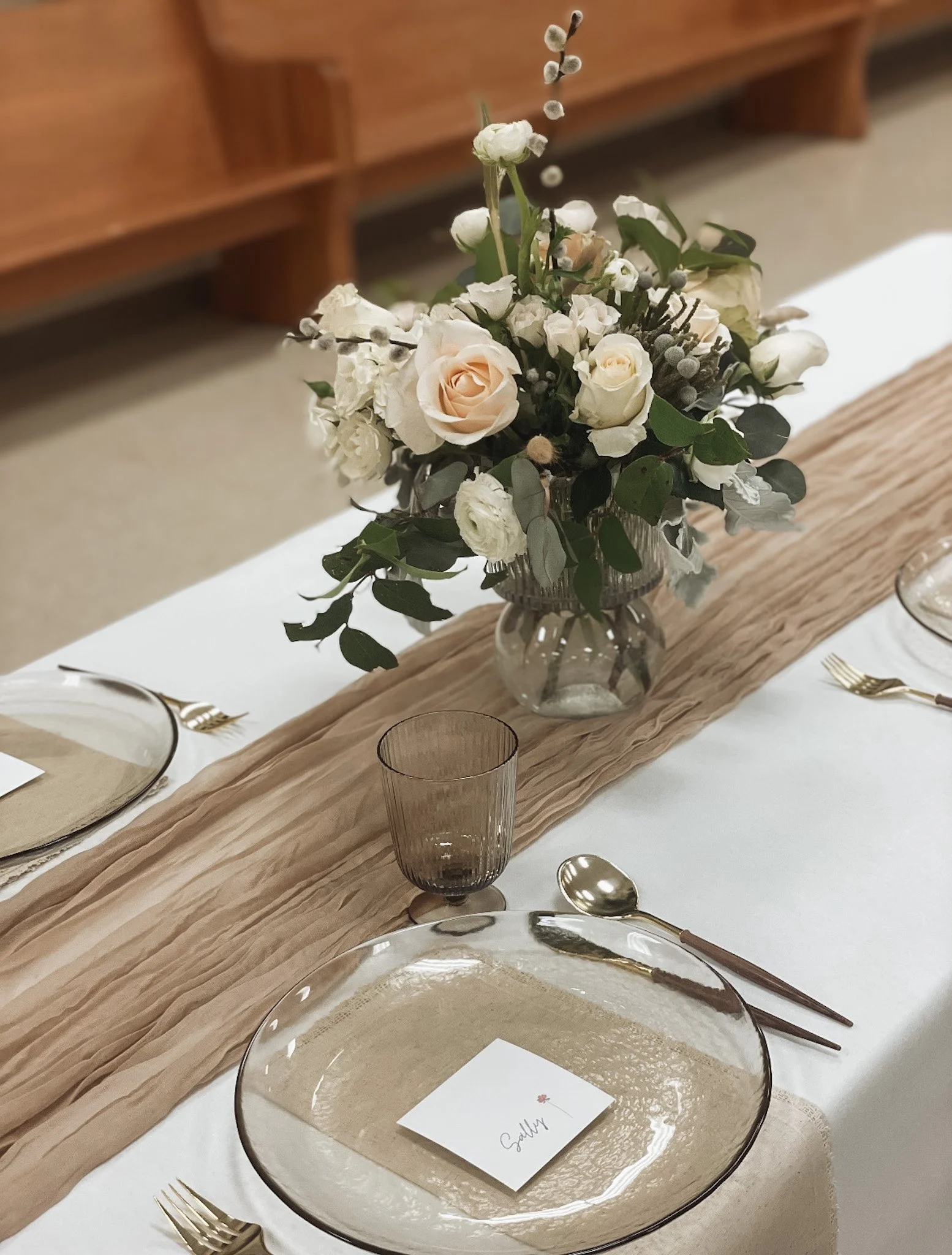 A table setting with a floral centerpiece in a glass vase, including white roses and greenery. The table has a beige runner with place settings that include clear glass plates, gold utensils, and a brown-tinted glass. A place card with the name "Gall