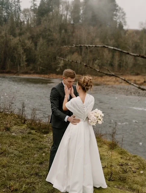 A bride and groom standing by a river in a romantic moment, with the bride holding a bouquet of white flowers and both dressed in wedding attire, surrounded by trees and a cloudy sky.
