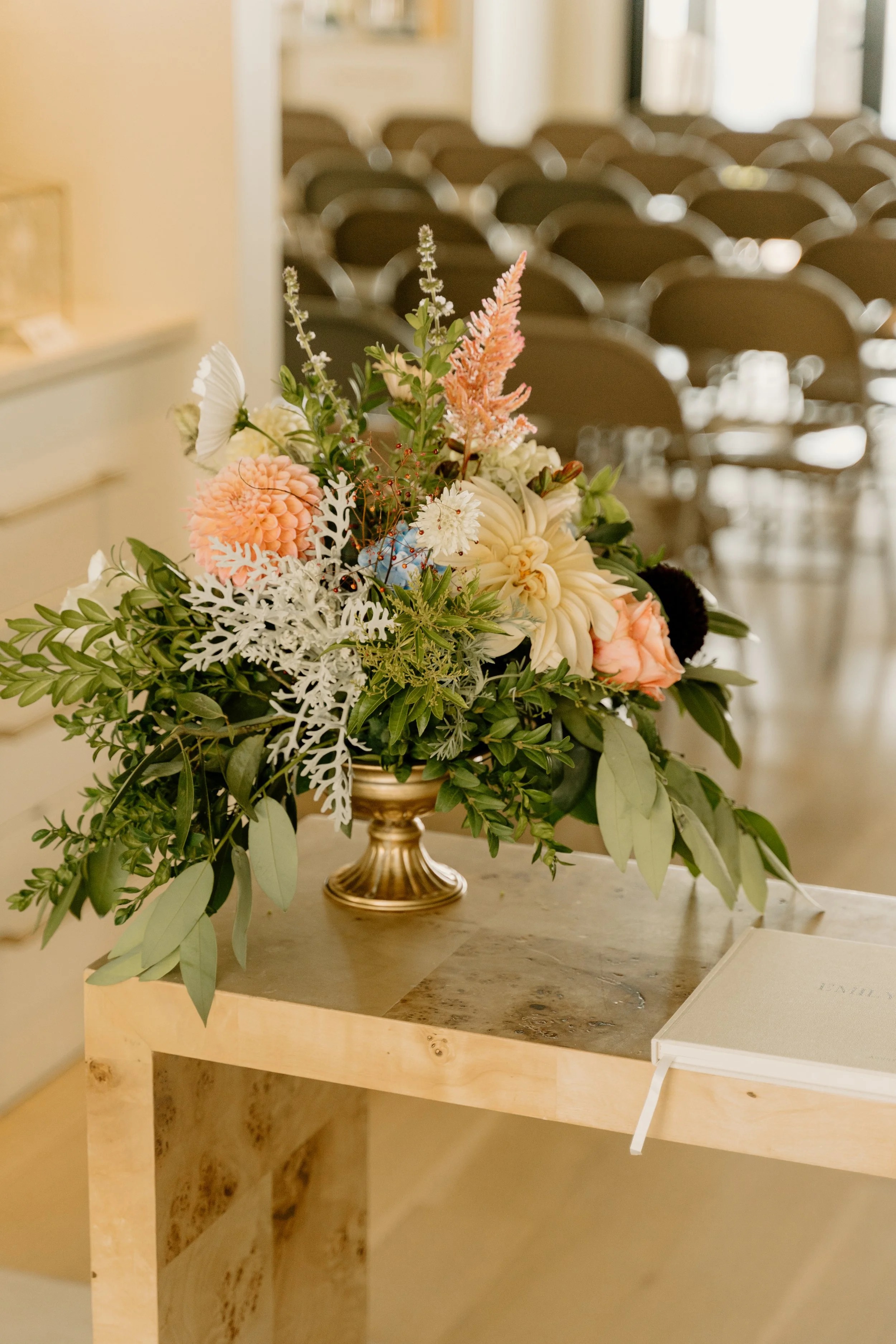 A floral arrangement featuring pink, white, and green flowers on a small wooden table, with a blurred background of brown chairs.