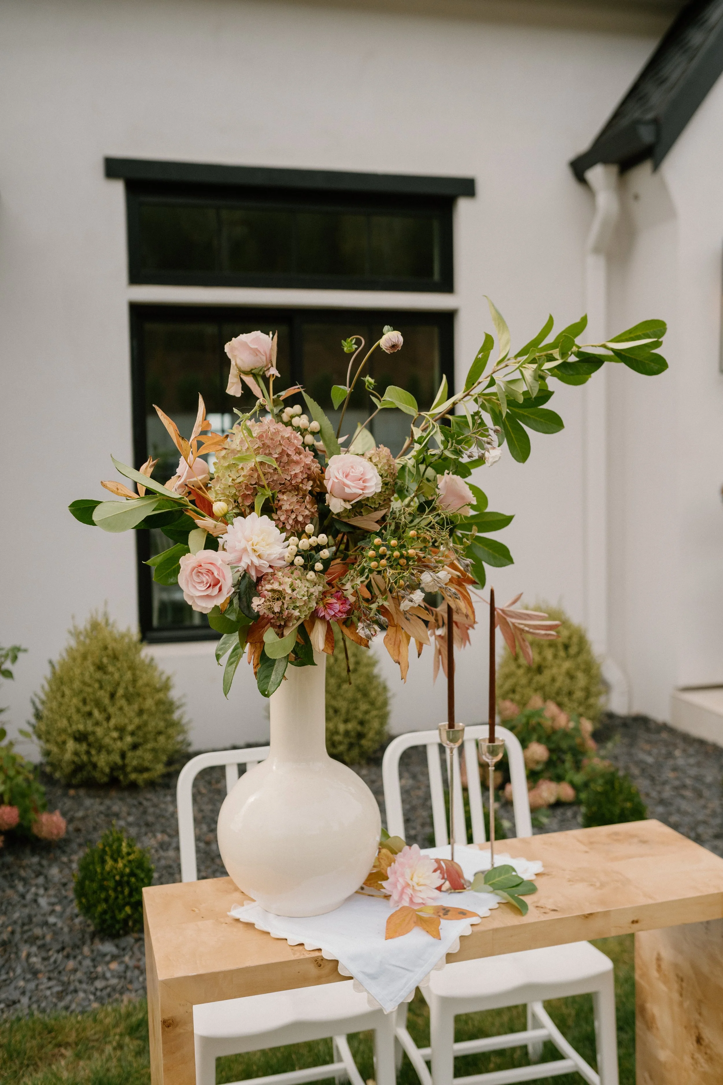 A large floral arrangement in a white vase on a wooden table outdoors, with two tall candles in holders, against a background of a white house with black windows and small green shrubs.