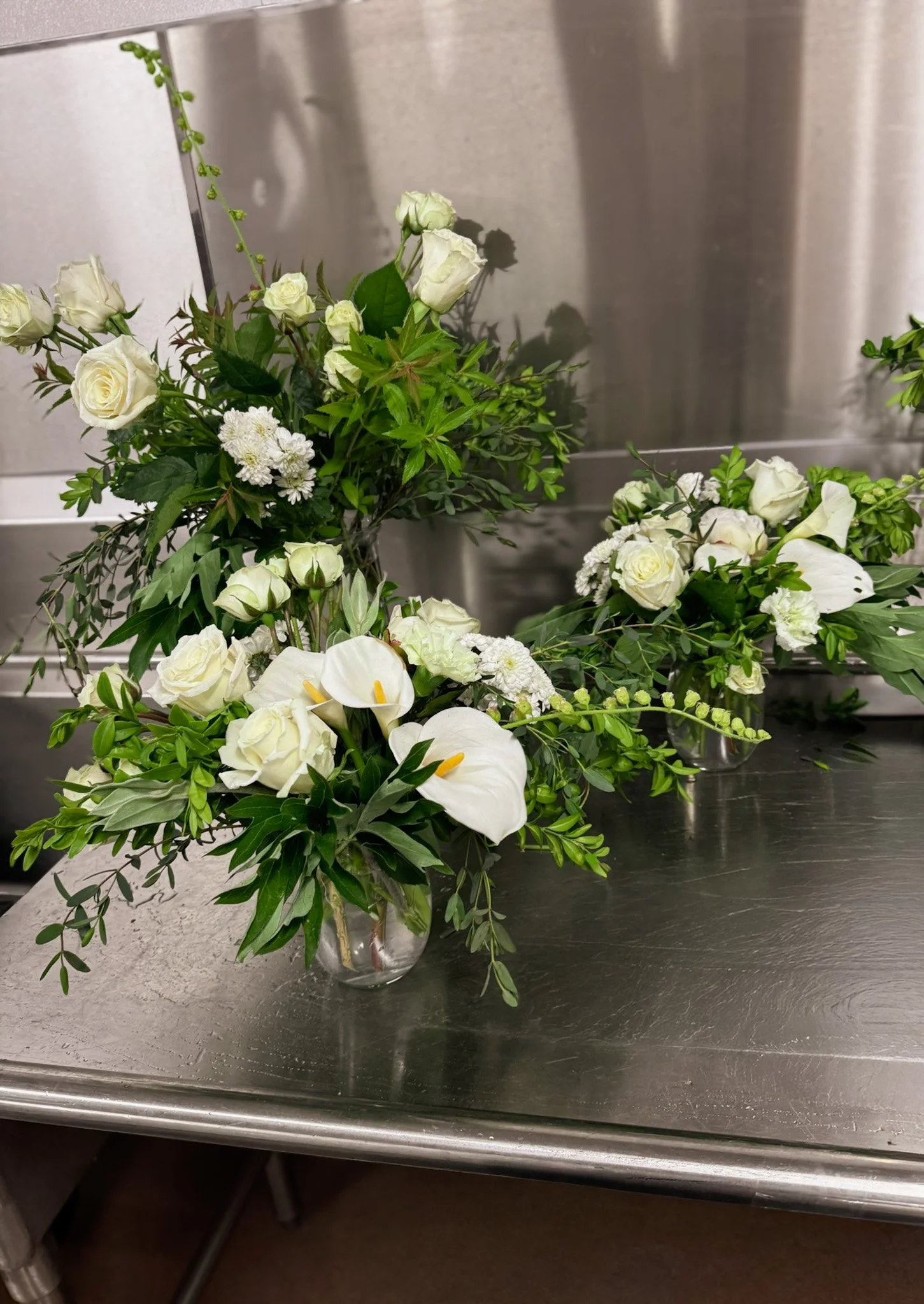 A floral arrangement with white roses, calla lilies, and other white flowers and green foliage in glass vases on a stainless steel table.