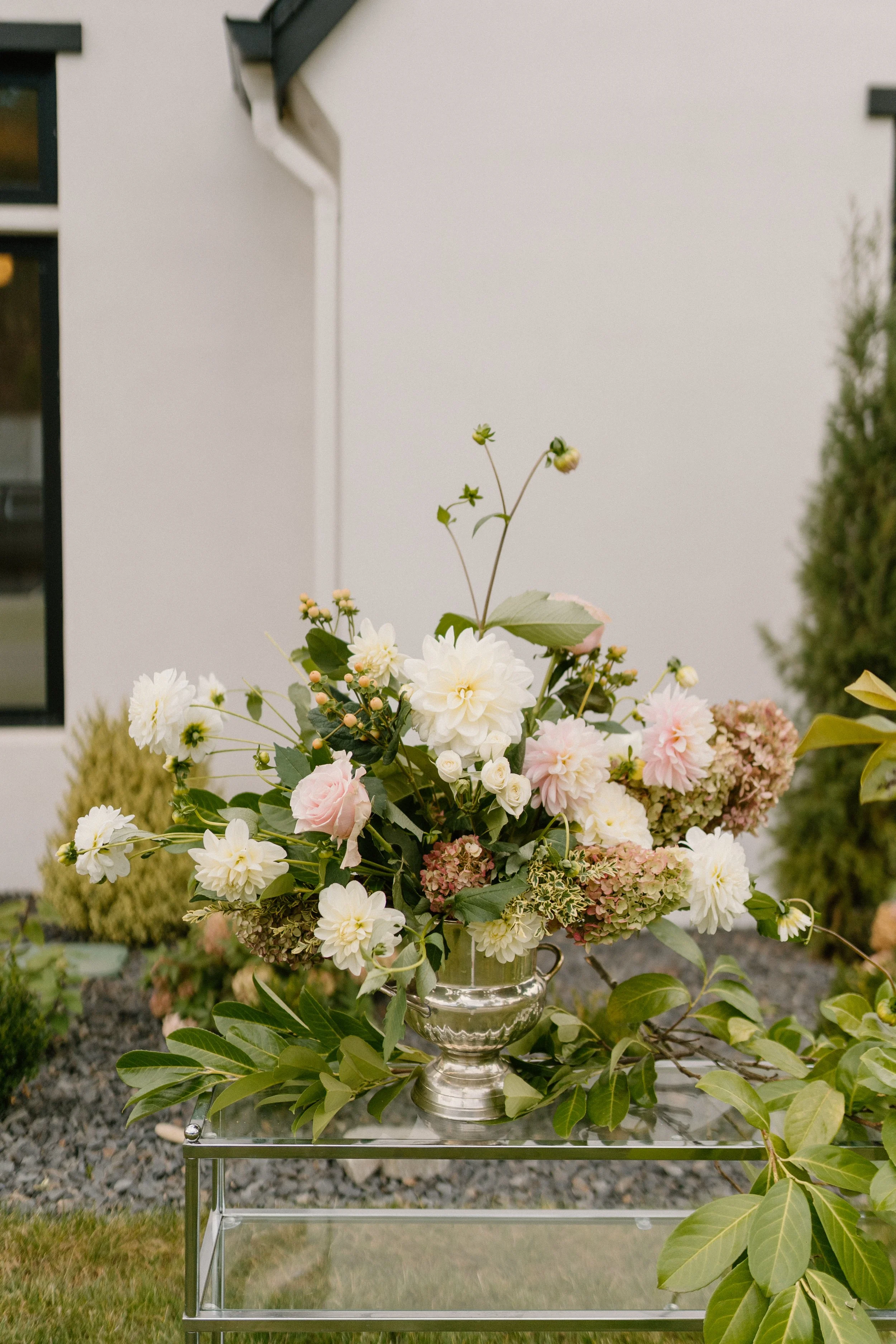 A floral arrangement with white and pink flowers in a silver vase placed on a transparent table outdoors.