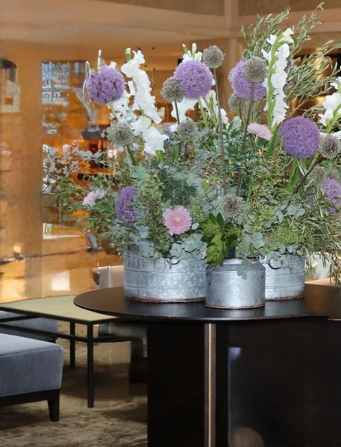 A large floral arrangement with purple allium, white gladiolus, and pink dahlias in galvanized metal containers on a black table.