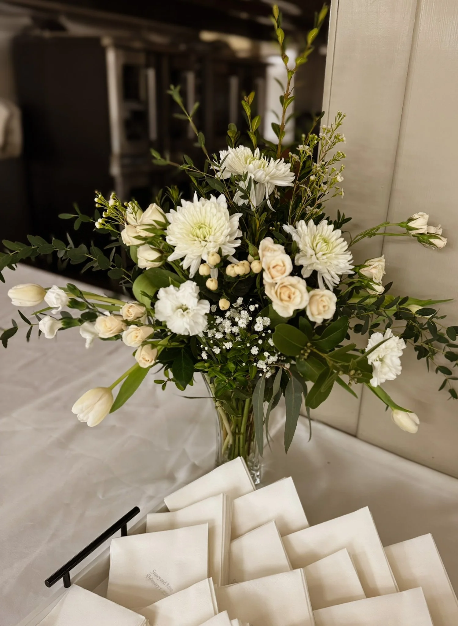 A bouquet of white flowers including chrysanthemums, roses, and tulips with greenery, placed on a table next to a stack of white napkins with text, in a vase