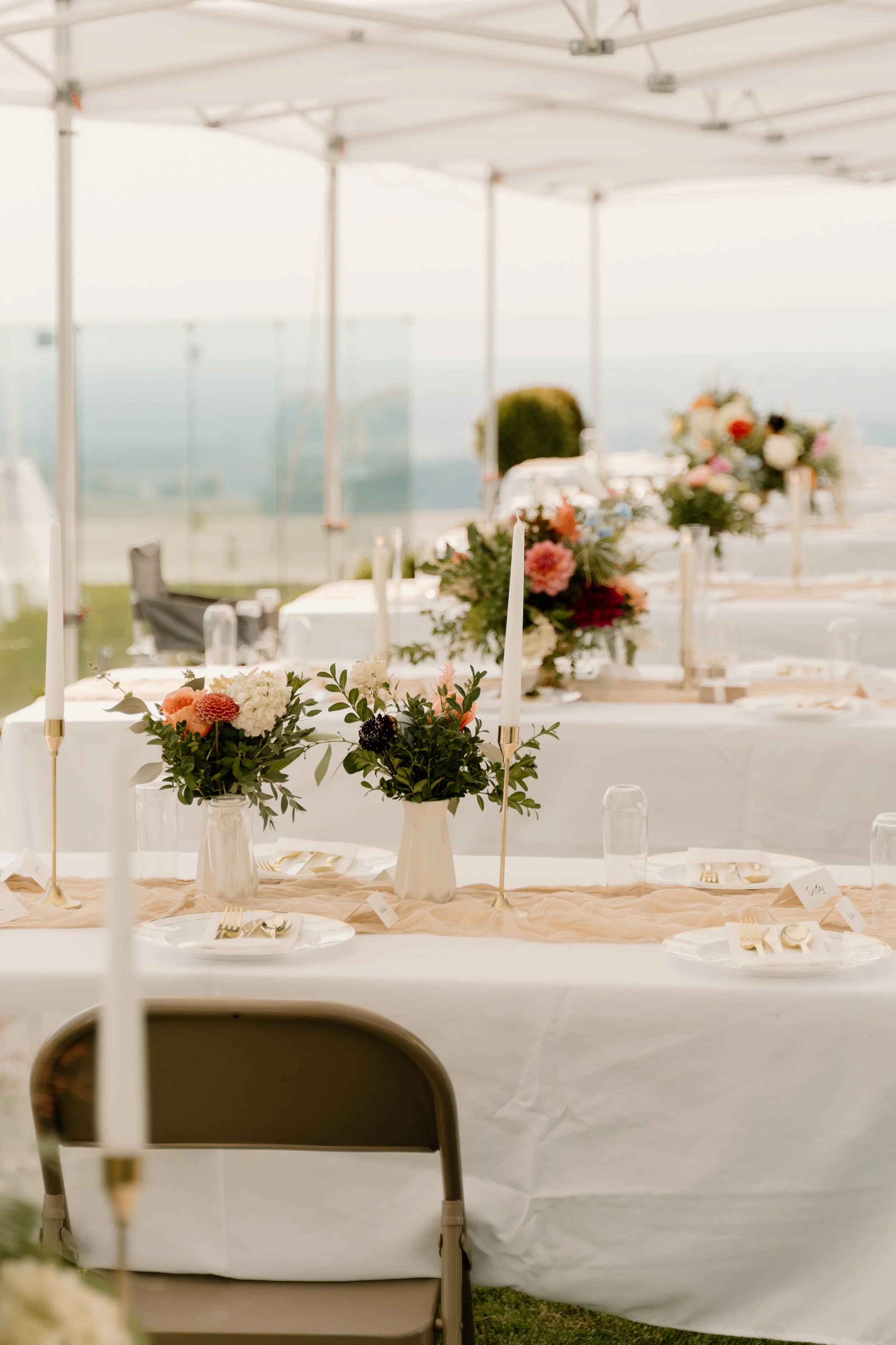 Elegant outdoor dining table decorated with pink and white floral centerpieces, tall candles, and white tableware, set under a white canopy with scenic view in the background.