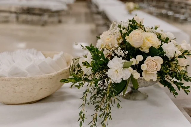 A floral arrangement with white and cream-colored roses and other white flowers, decorated with green leaves, on a white surface.