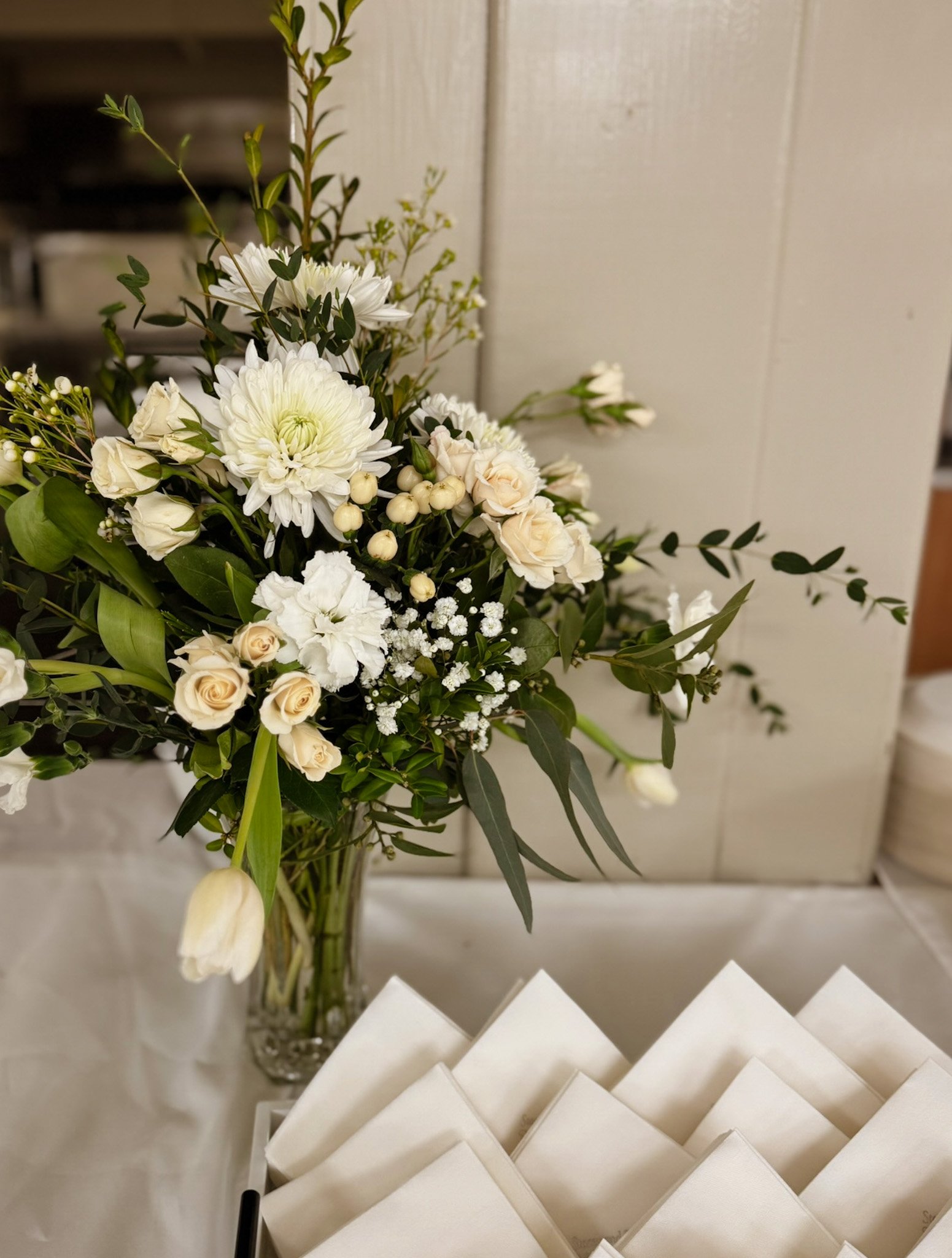 A floral arrangement with white flowers and green foliage in a glass vase on a table with white napkins.