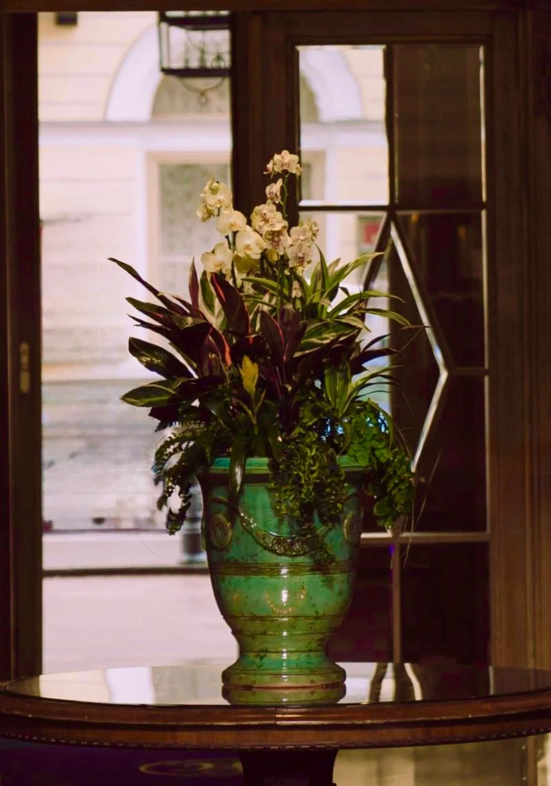 A green ceramic vase with a floral pattern contains a bouquet of white orchids and green leafy plants, placed on a polished wooden table inside.