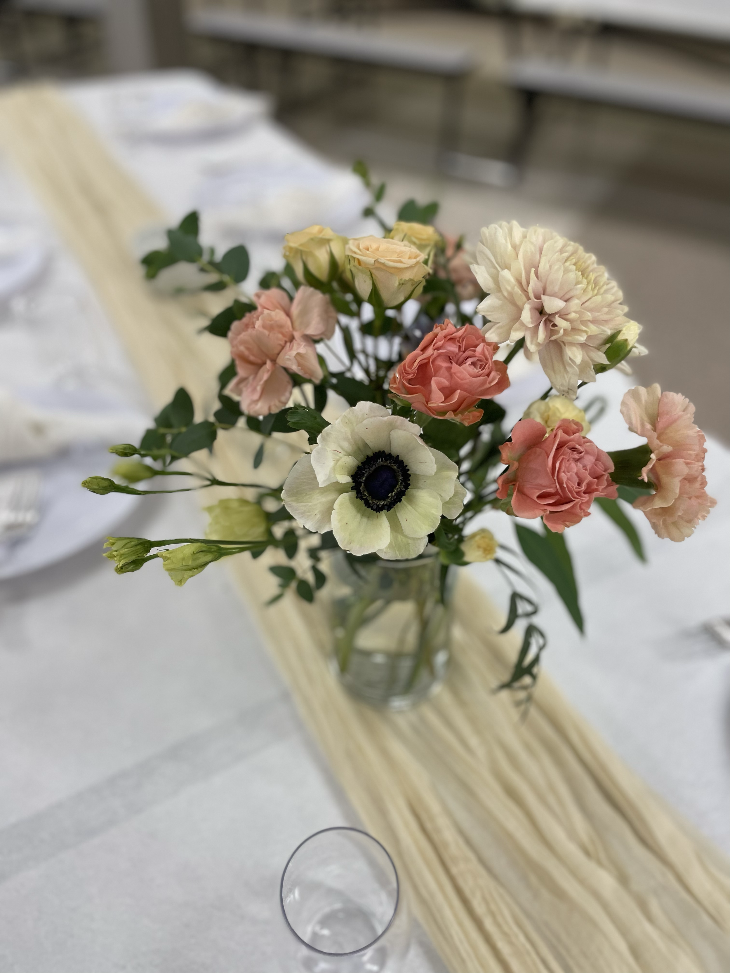 A bouquet of pastel-colored flowers in a glass vase on a table with a white tablecloth and a beige table runner.