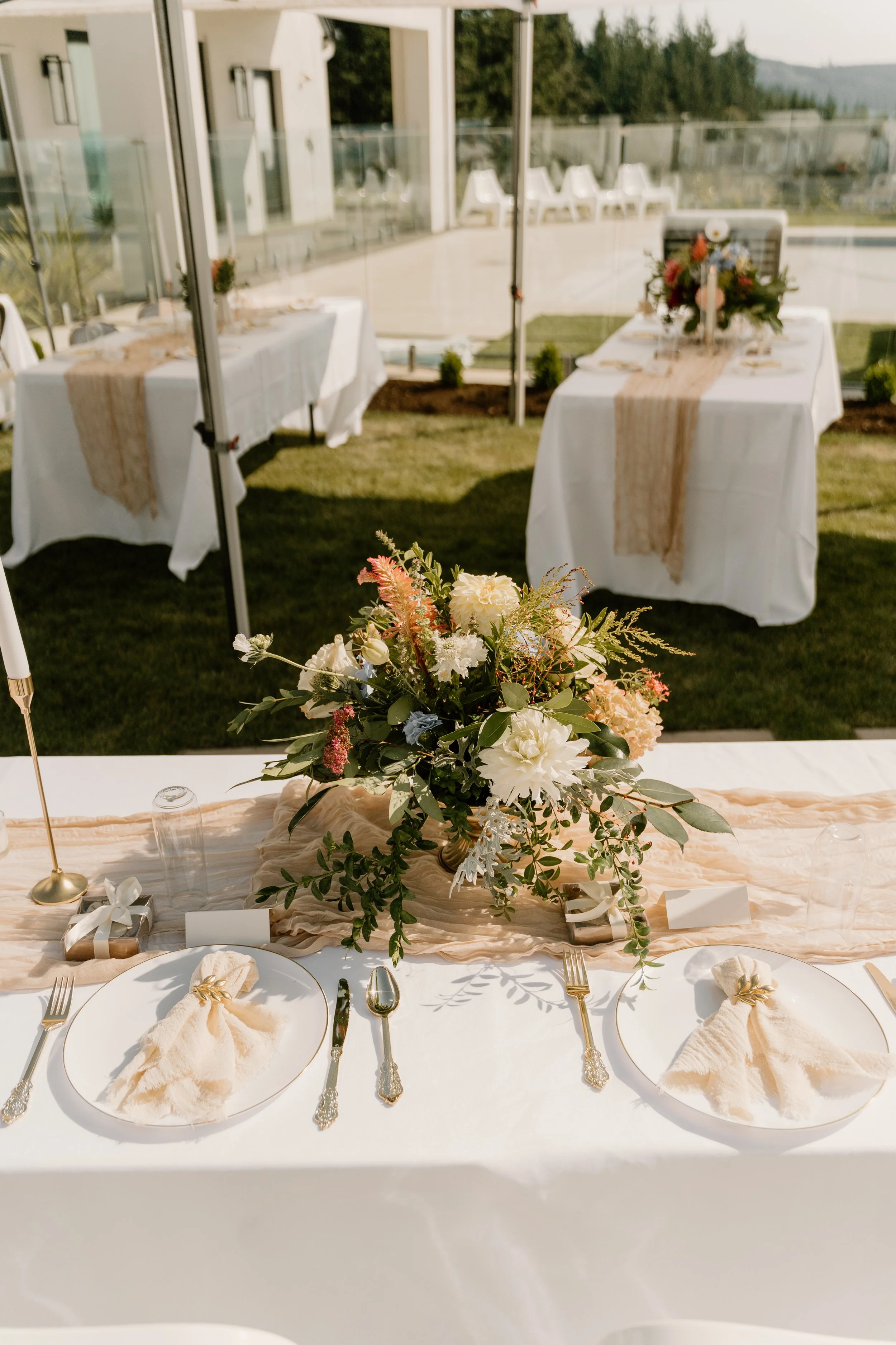 Elegant outdoor dining setup with a floral centerpiece, arranged for a special occasion like a wedding, featuring two place settings with white plates, cloth napkins, gold utensils, and glassware, on a table draped with a peach-colored runner. In the