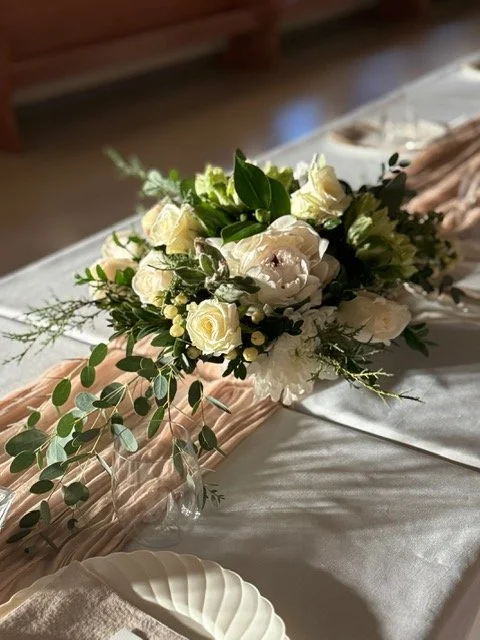 A floral arrangement with white roses, green leaves, and greenery on a table decoratively arranged with a light pink fabric and a white tablecloth.