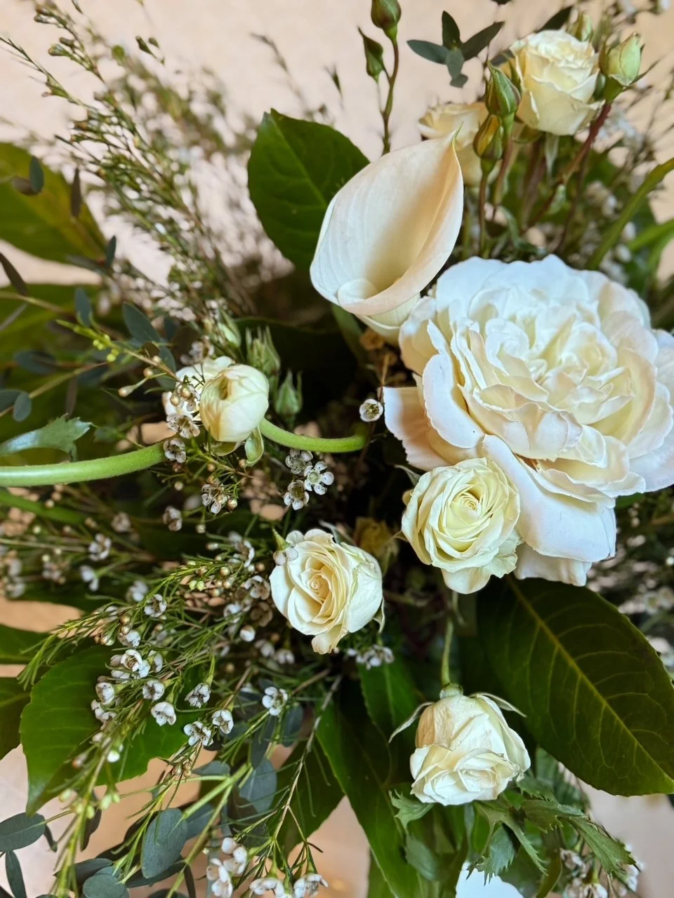 A bouquet of white flowers, including roses, calla lilies, and small filler flowers with green leaves.