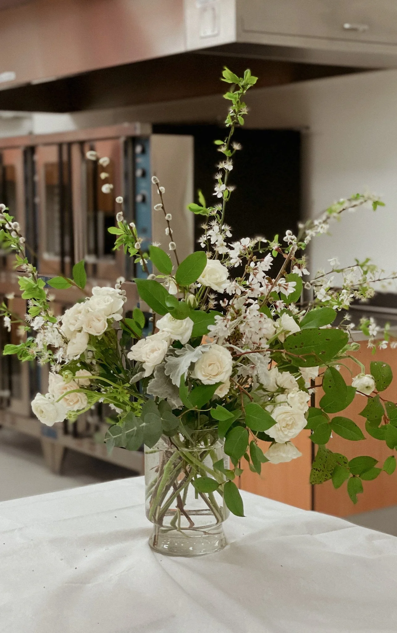 A glass vase with white roses, small white blossoms, and green leaves on a white tablecloth, with a blurred office background.
