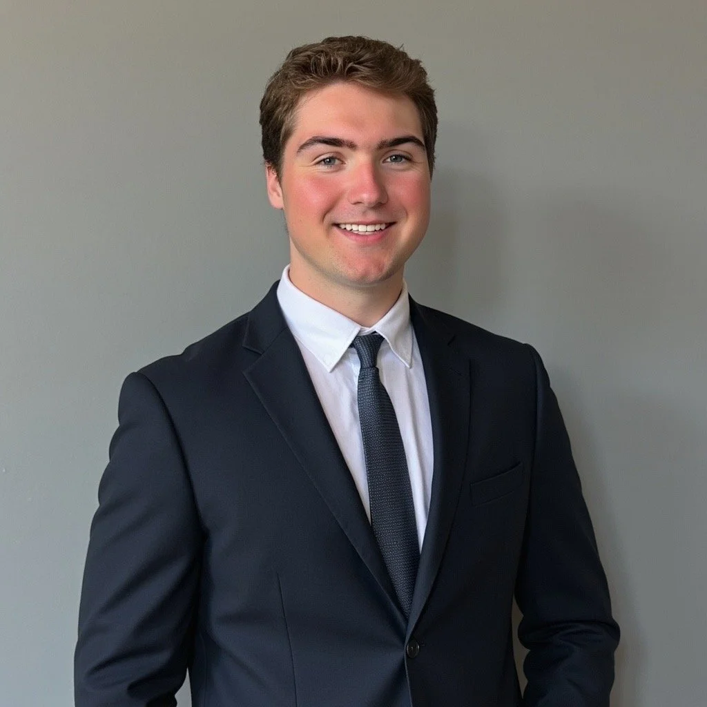 A young man in a dark suit, white shirt, and black tie, smiling and posing against a light gray wall.