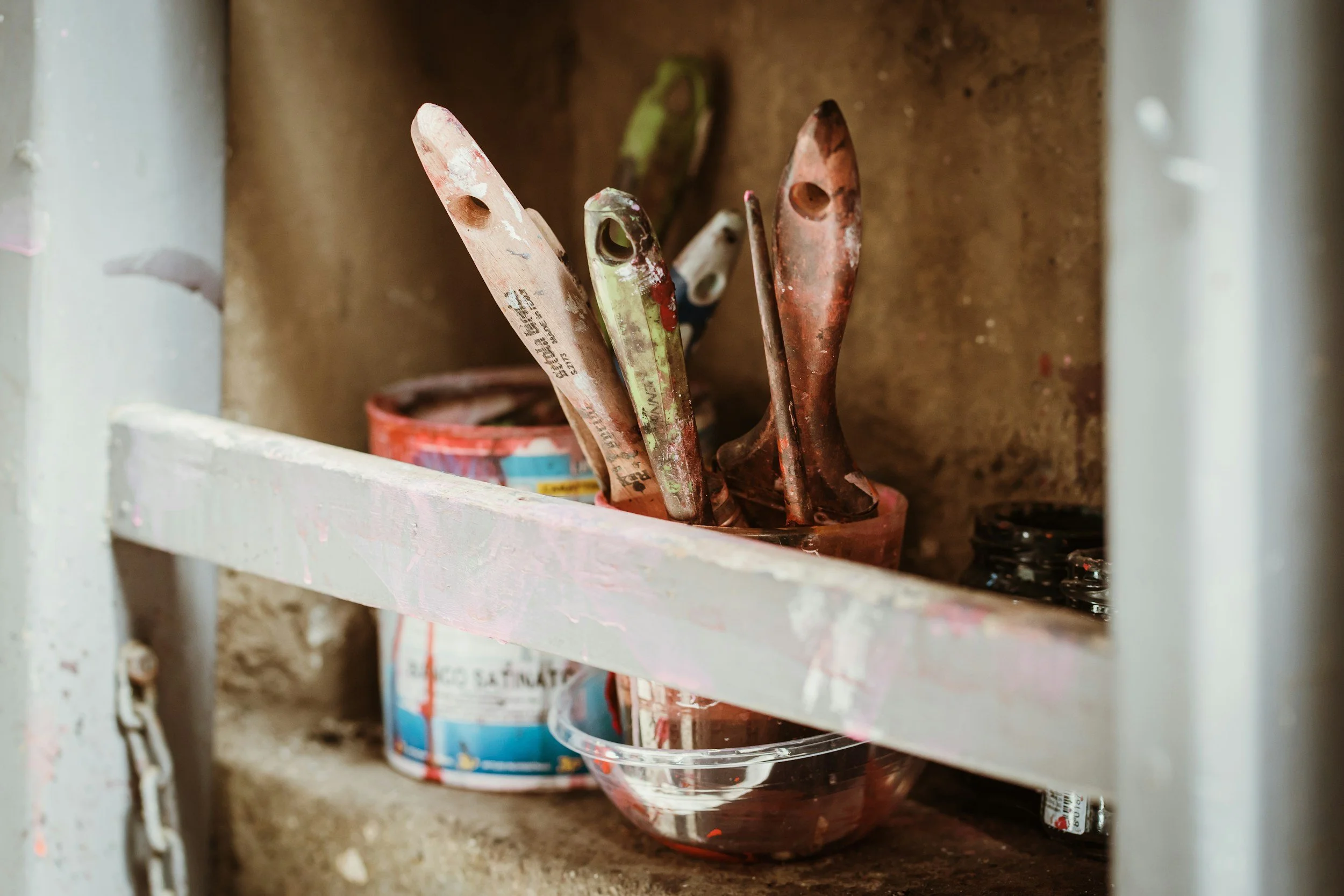 Paintbrushes with dried paint in a glass bowl on a dusty shelf, with paint cans behind them.