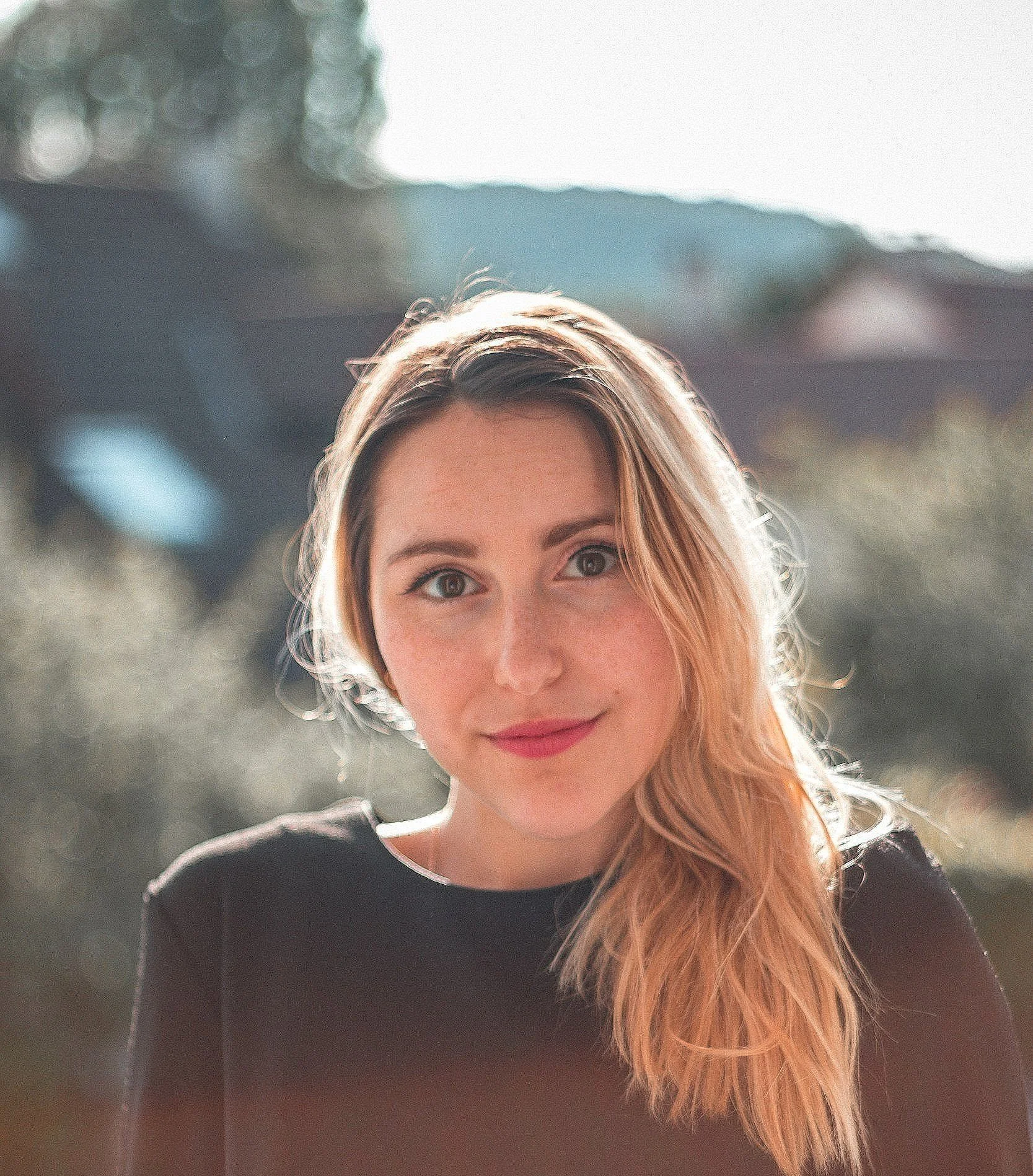 A young woman with long reddish-blonde hair, wearing a black top, smiling outdoors with a blurred background of trees and sky.