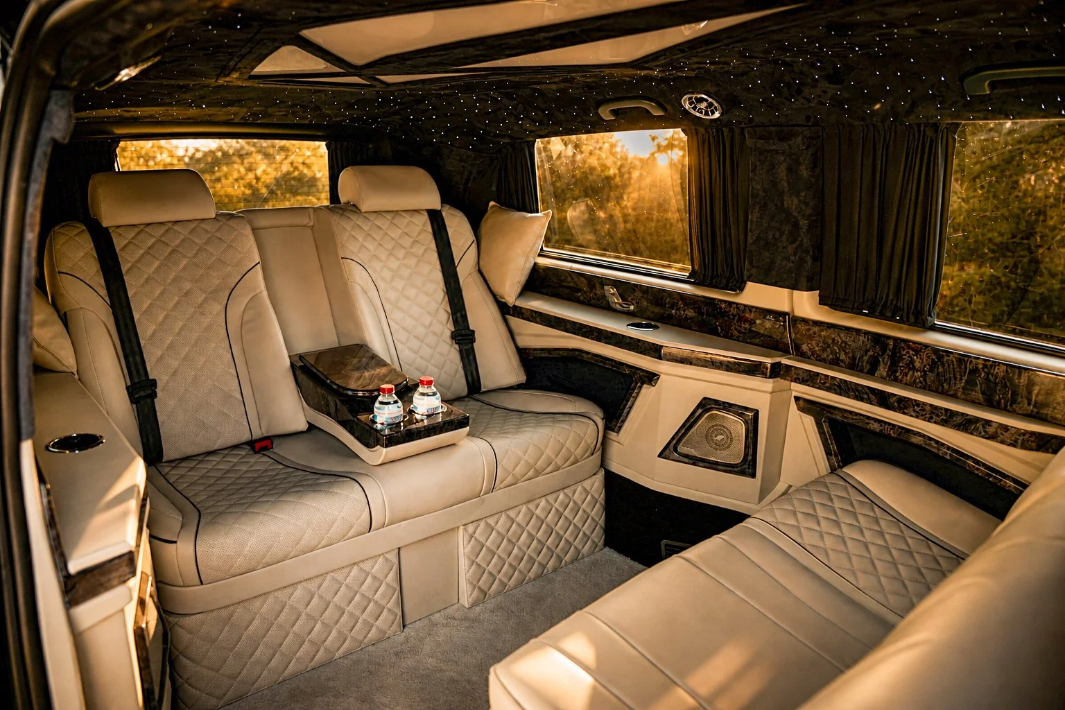 Interior of a luxurious van with beige quilted leather seats, a small table with water bottles, black curtains, and star-like lights on the ceiling during sunset.