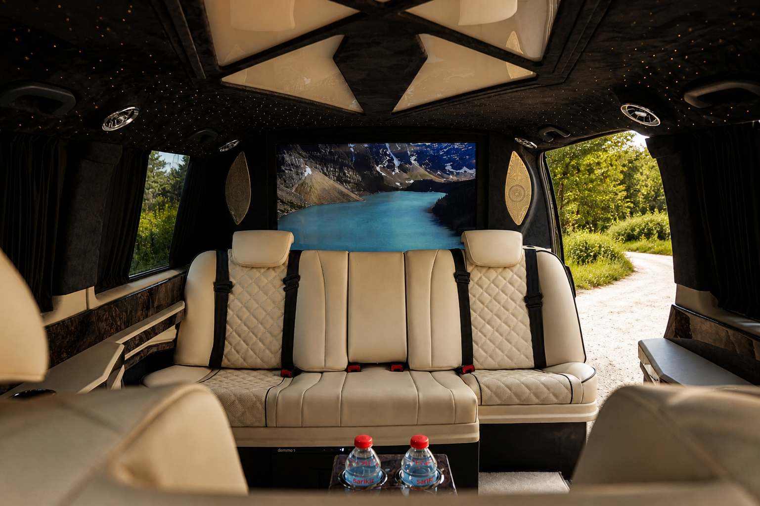 Interior of a luxury van with cream-colored leather seats, a scenic mountain lake view on a screen, and bottles of water on a table, with open doors revealing a green outdoor landscape.
