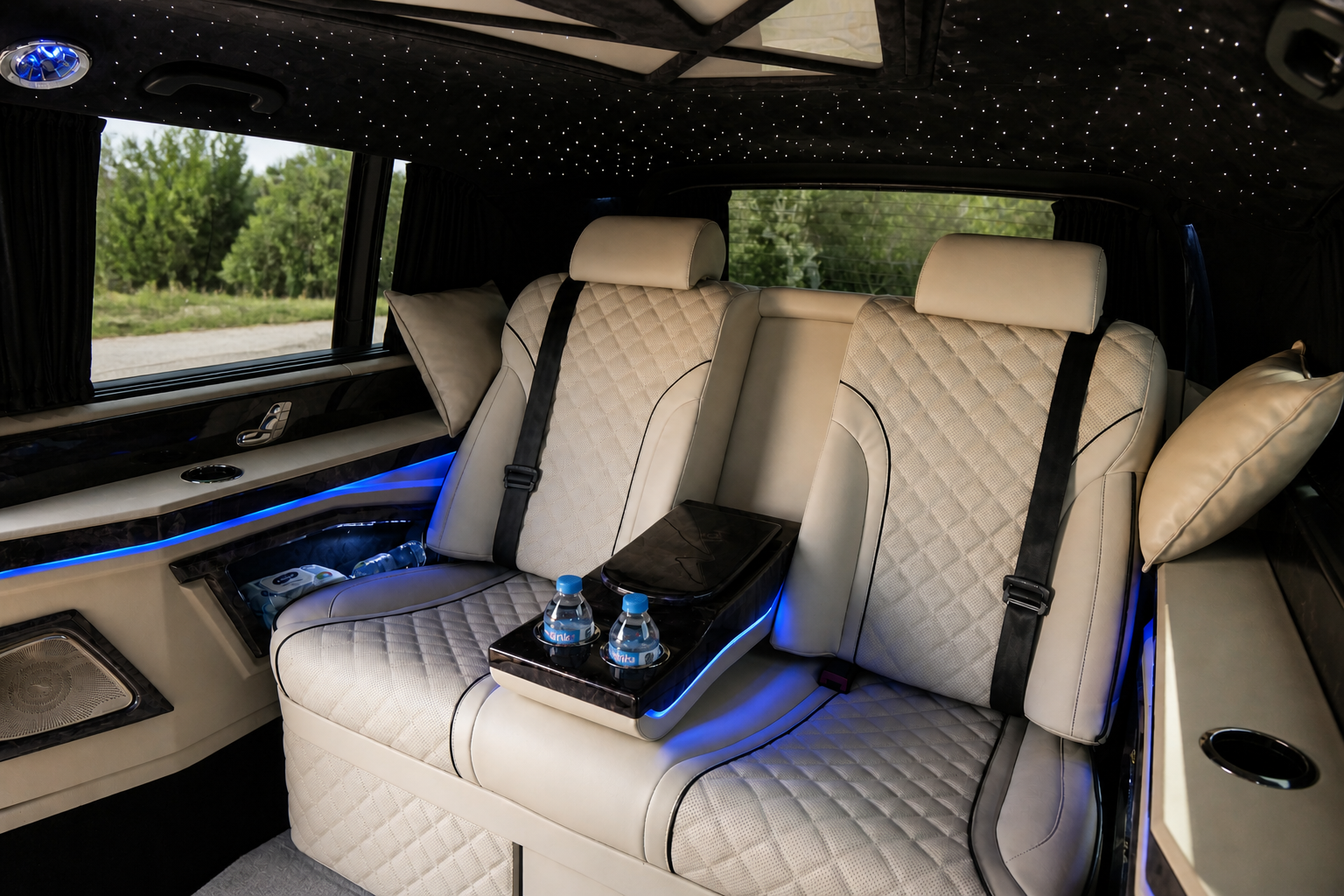 Luxury interior of a limousine with beige quilted seats, headrests, pillows, and black seat belts, illuminated with blue LED lighting, with windows showing greenery outside and small star-like lights on the ceiling.