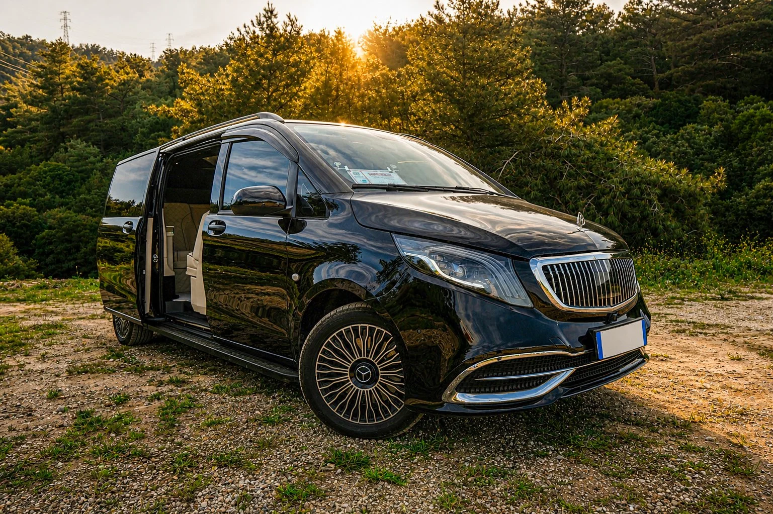 Black Mercedes-Benz van with open sliding door parked on gravel with green foliage and trees in the background during sunset.
