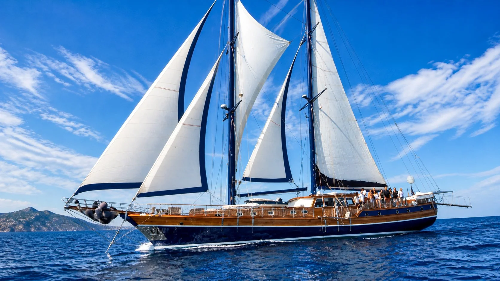 A large sailing yacht with white and dark sails on the ocean under a partly cloudy sky, with a group of people on deck and a distant landmass visible on the horizon.