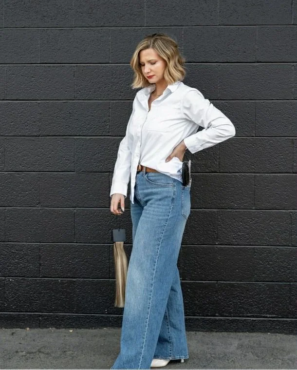 Woman standing against a black brick wall, wearing a white shirt and blue jeans, holding a hair extension bundle.