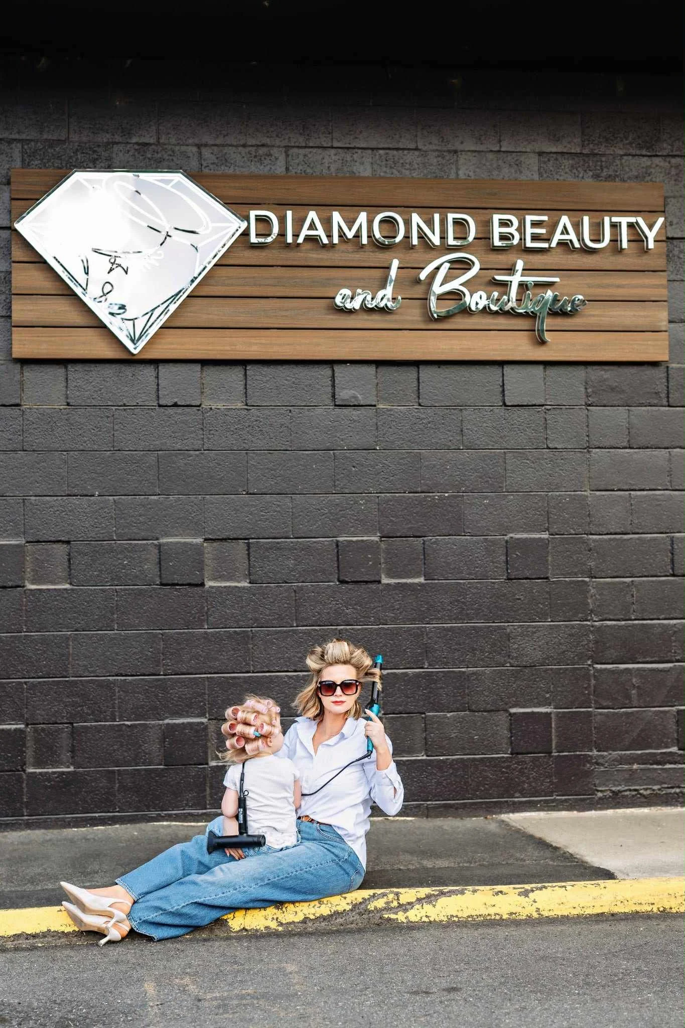 A woman with blonde hair, sunglasses, and a white shirt sitting on the street curb, holding a young girl with curly hair, hair rollers, and a white shirt. They are in front of a black brick wall with a store sign that says 'Diamond Beauty and Boutique' with a diamond logo in white.