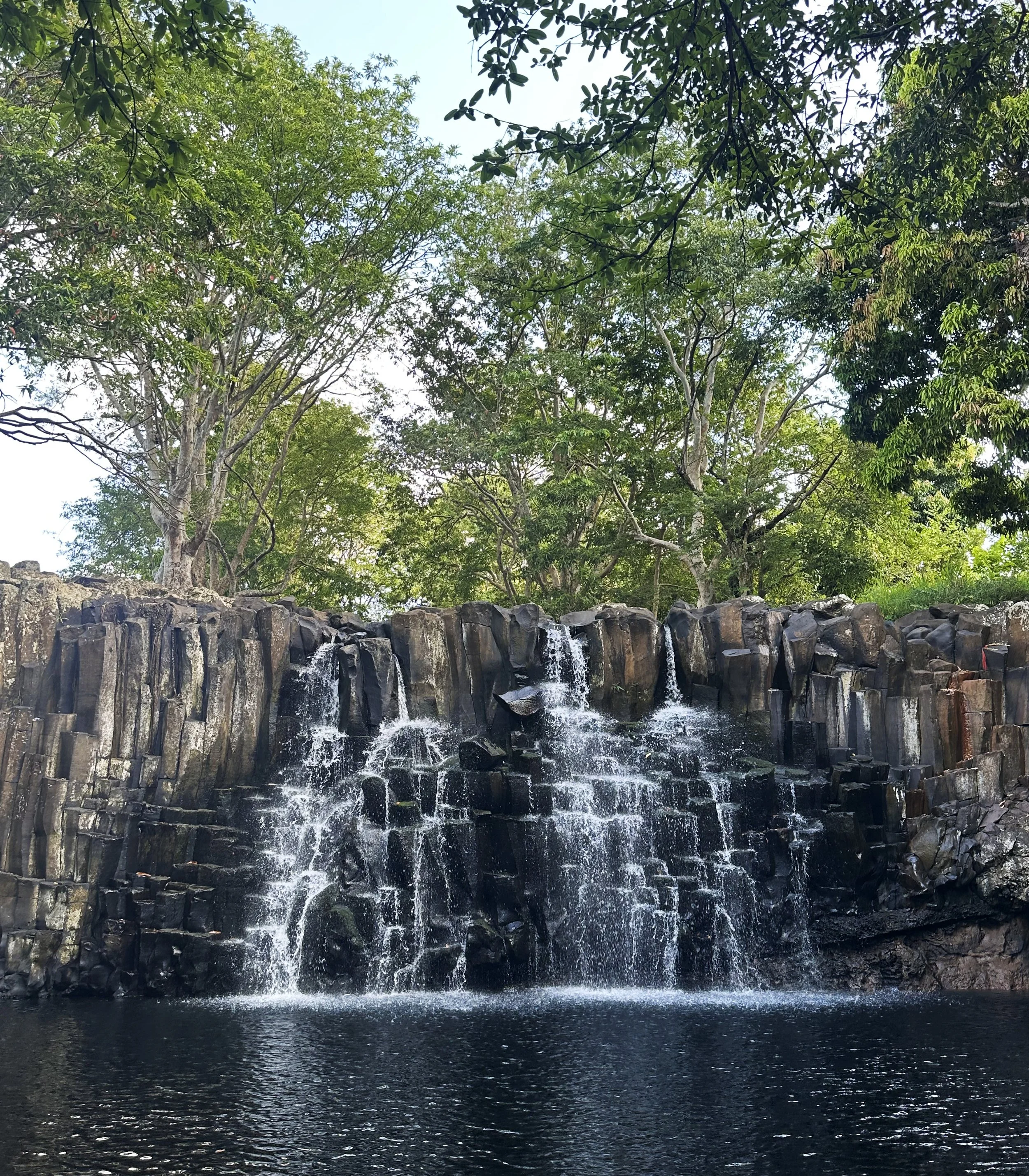 A waterfall cascading over dark rocks surrounded by green trees, with a clear sky visible above in Mauritius