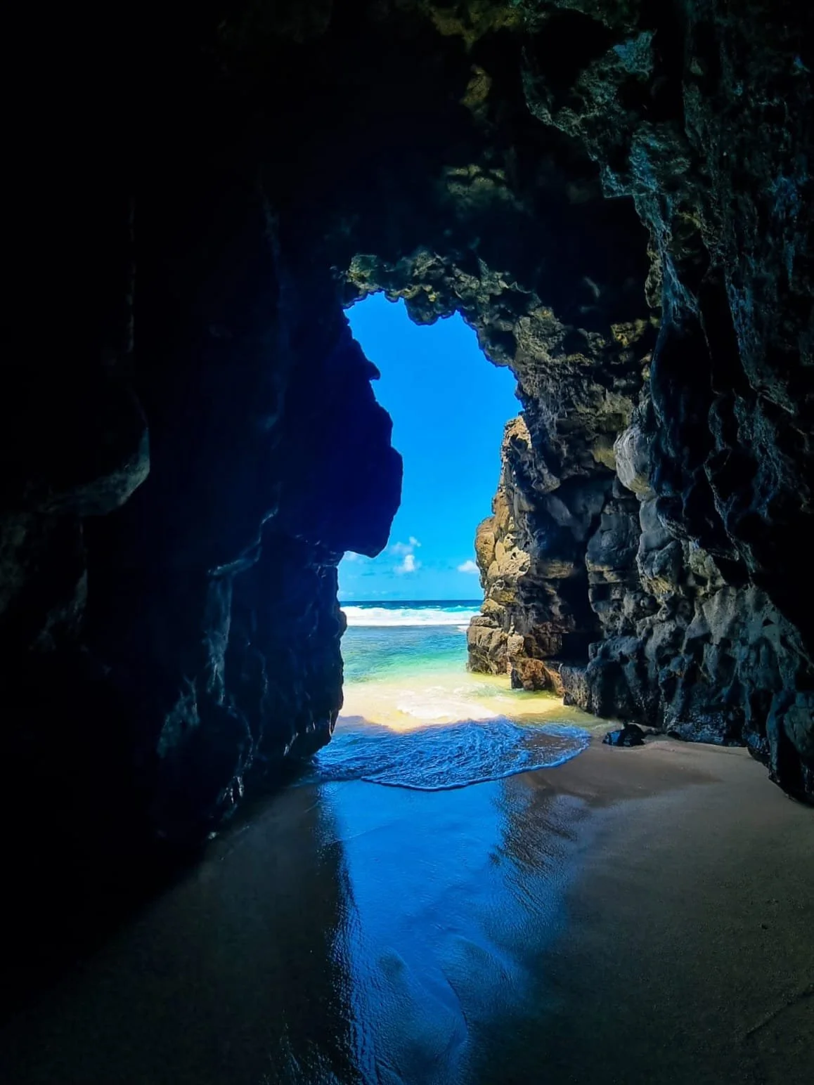 View of the ocean and sandy beach seen through a rocky cave opening with blue sky and white clouds in the background in Mauritius.