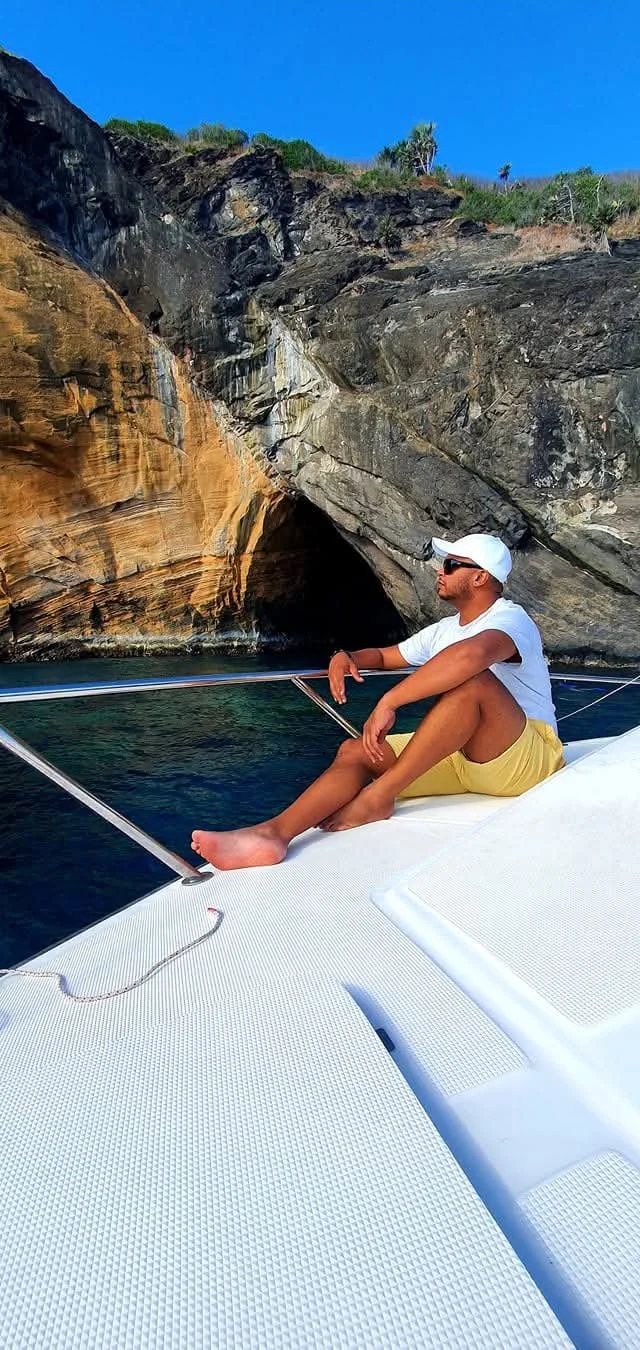 A man sitting on the deck of a boat near a rocky cliff, wearing a white cap, sunglasses, a white T-shirt, and yellow shorts, with water and a clear blue sky in the background in Mauritius.