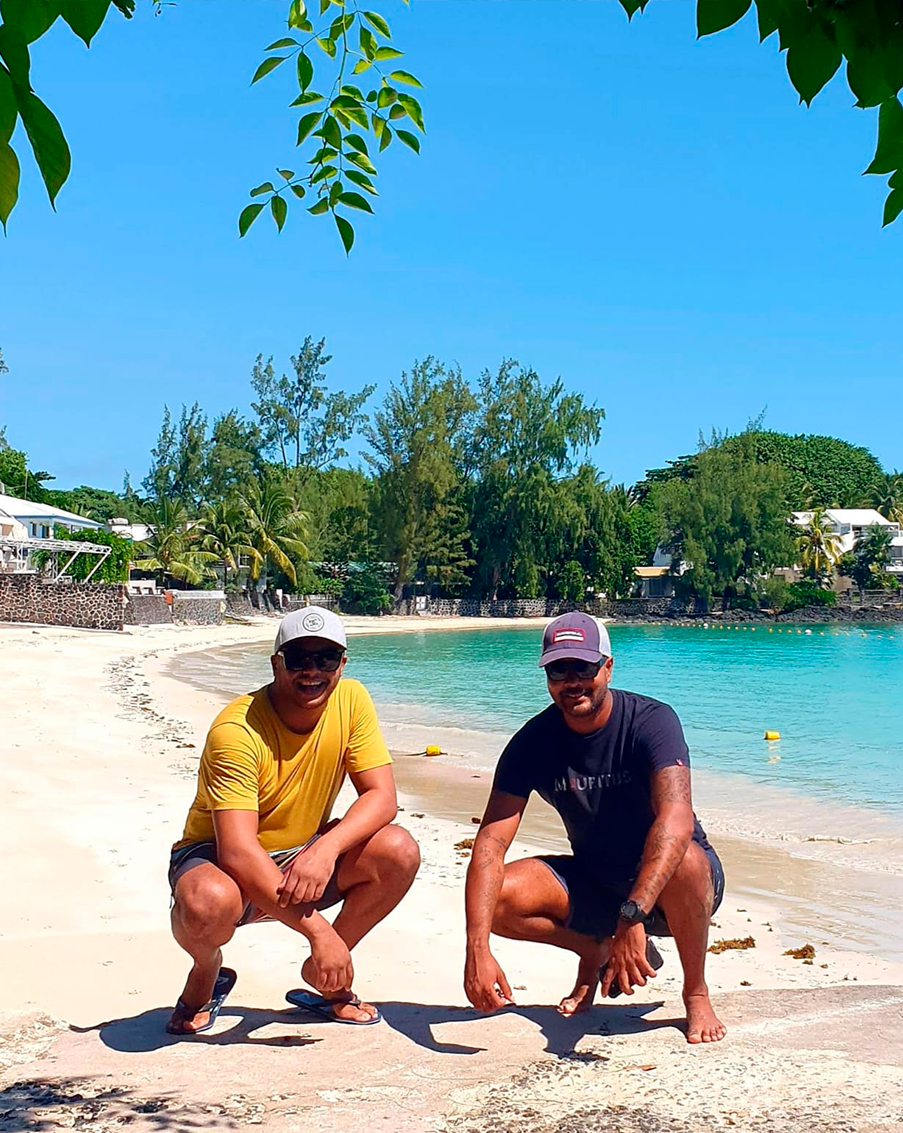 Two men squatting on a sandy beach near the water, smiling at the camera. They are wearing casual summer clothing and caps, with trees and buildings in the background under a bright blue sky in Mauritius.