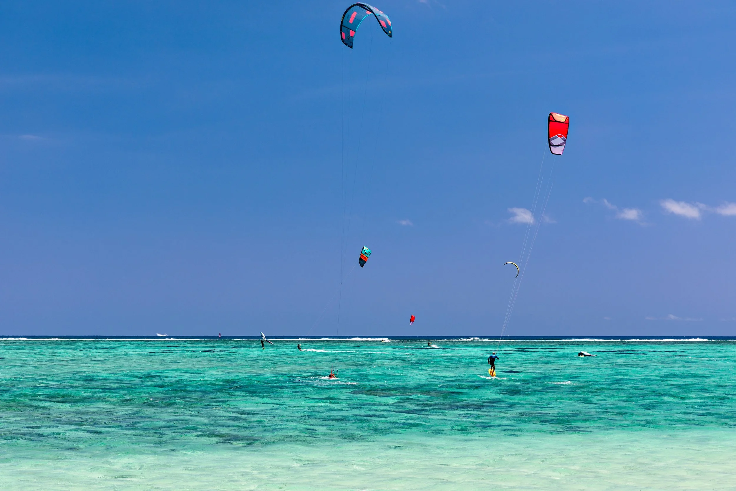People kite surfing in the ocean with clear turquoise water under a blue sky, with a few other kite surfers and boats visible in the distance.