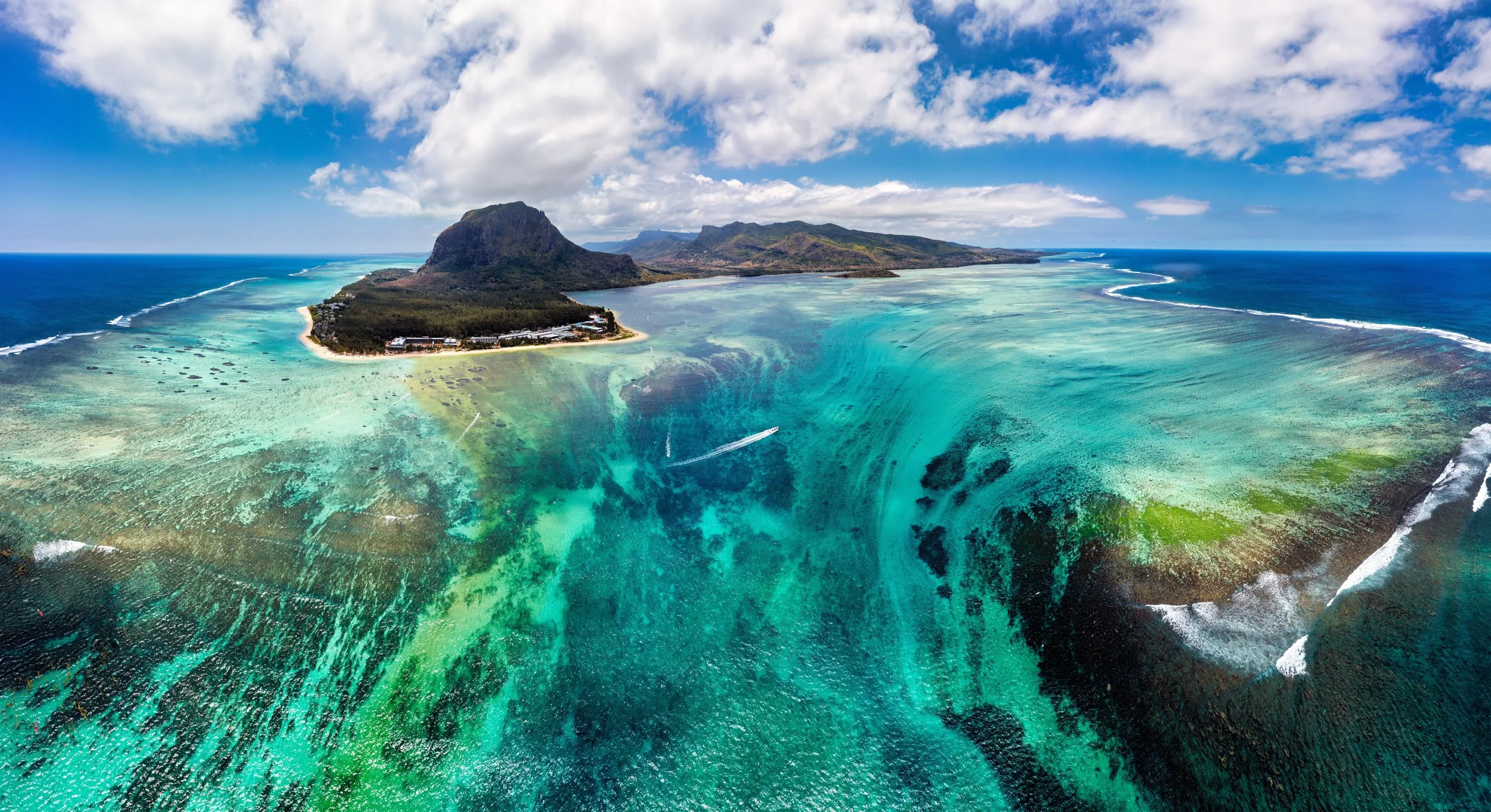 Aerial view of a tropical island with a mountain, surrounded by turquoise waters, white sand beaches, and coral reefs, under a partly cloudy sky.