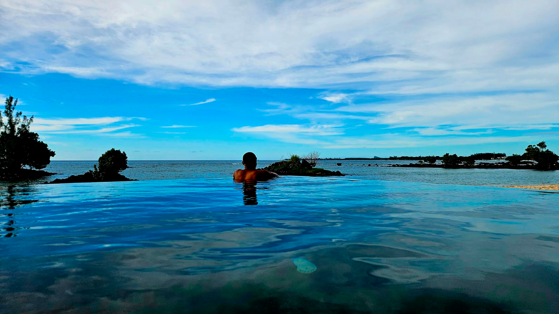 Person lounging in an infinity pool overlooking a tropical coastline with trees and the ocean, under a partly cloudy sky.