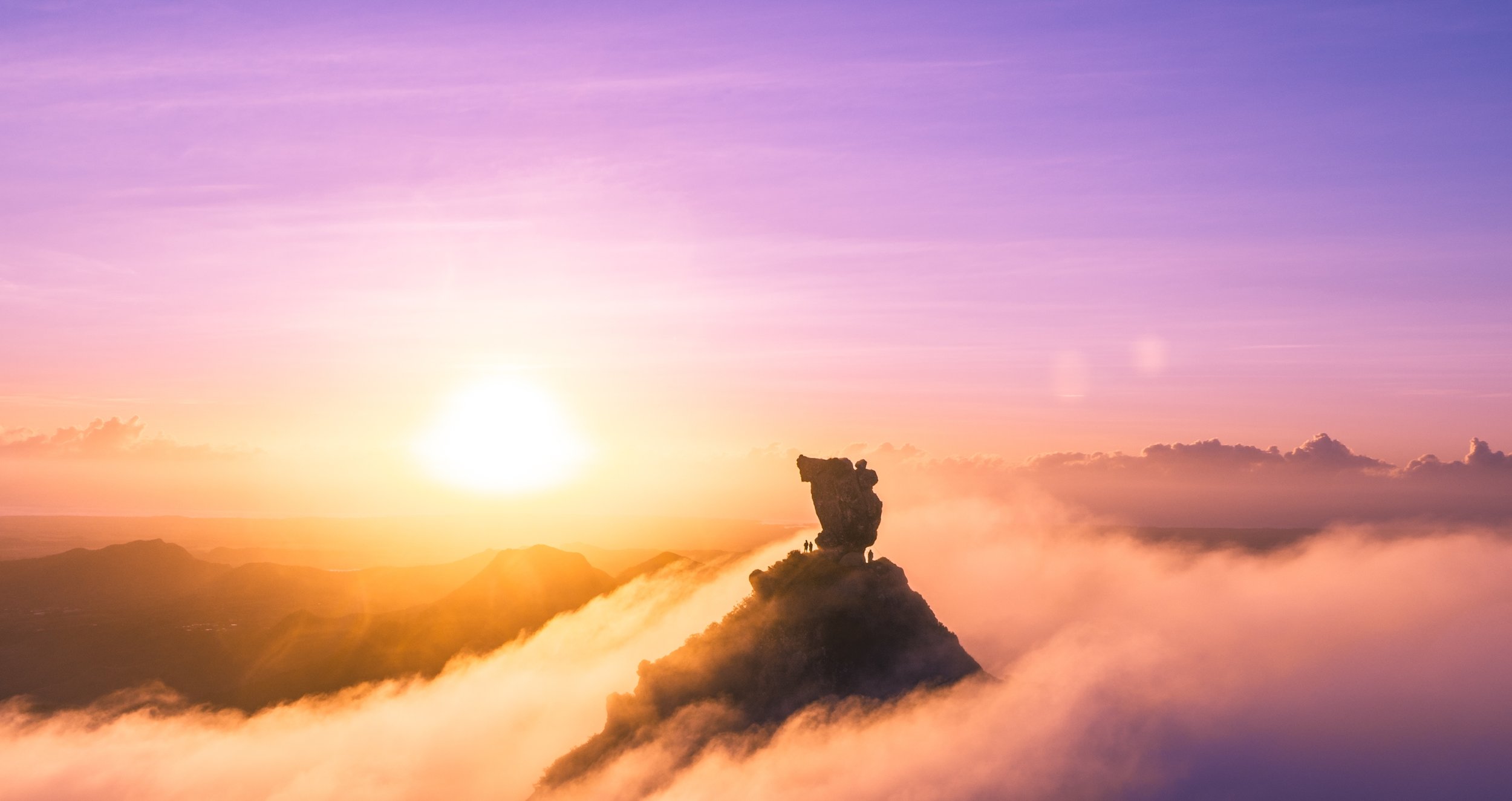 A scenic sunset over mountains with a unique rock formation in the foreground, wispy clouds, and a colorful sky in shades of purple, pink, and orange.
