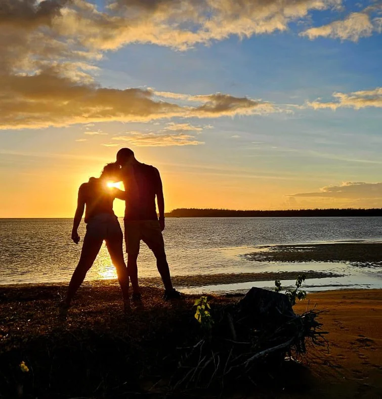 A couple standing on a beach during sunset, with their heads touching and arms around each other, silhouetted against the colorful sky and water in Mauritius.