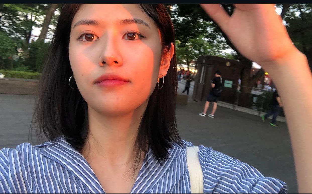 A young woman with black hair and hoop earrings taking a selfie outdoors at sunset, with trees and other people in the background.