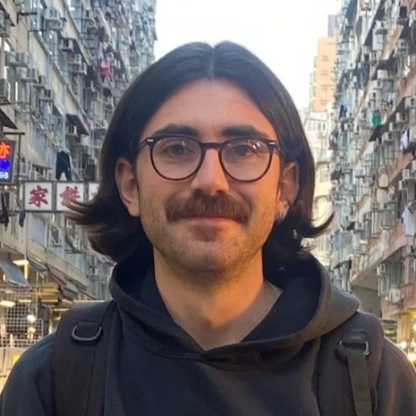 Young man with glasses and a mustache smiling at camera on a busy city street with tall buildings and hanging laundry.