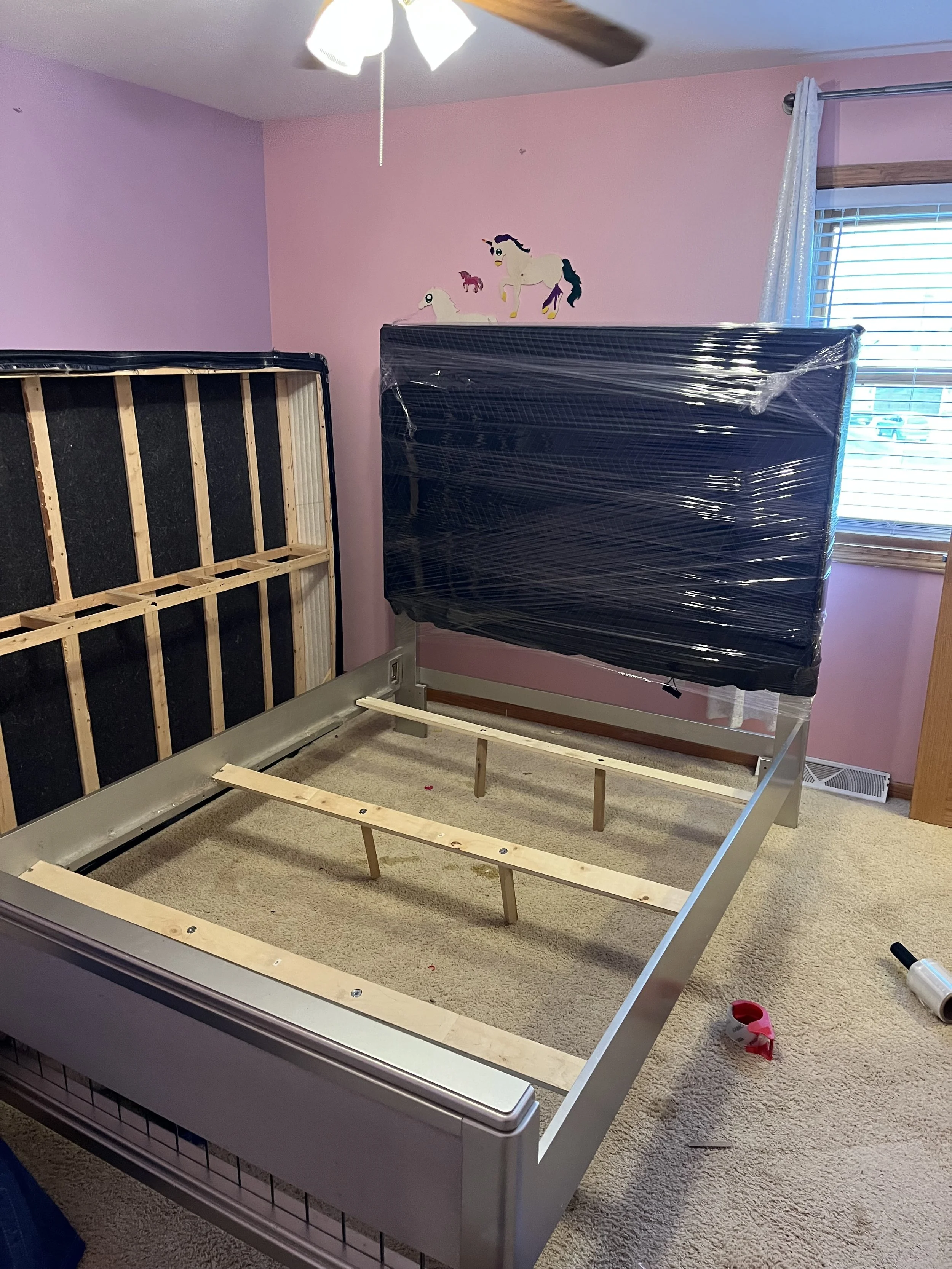 Room with pink walls, a window with blinds, a ceiling fan, and a partially assembled bed frame under plastic. There is a wall decal of unicorns and a small horse. The carpet is beige and there are construction supplies on the floor.