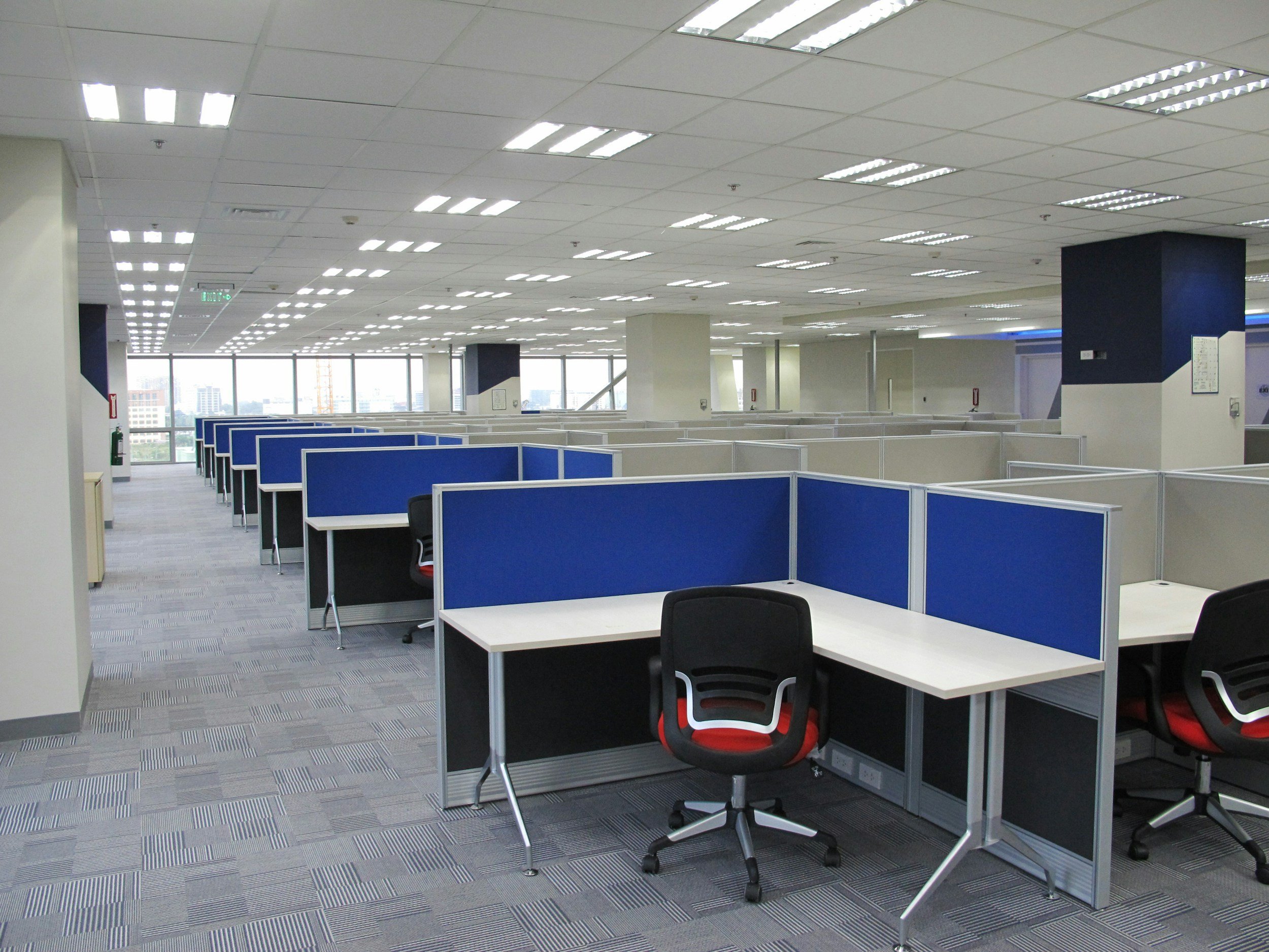 An empty office space with numerous cubicles with blue partitions, chairs, and a view of city buildings through large windows. The ceiling has many fluorescent lights.