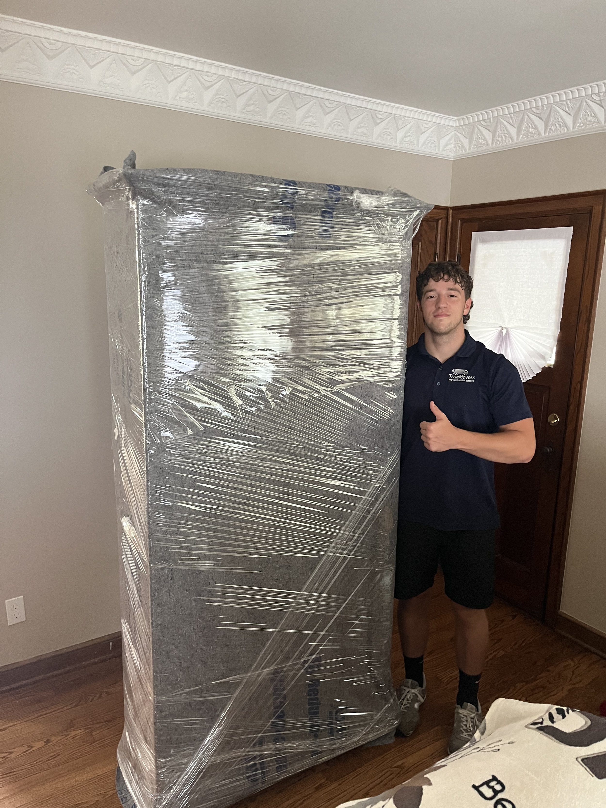 A young man is standing indoors next to a large piece of furniture wrapped in plastic, giving a thumbs up and smiling. He is wearing a navy blue polo shirt, black shorts, gray sneakers, and socks. There is a closed door, a decorative white fan on the window, and a beige wall with white crown molding in the background.