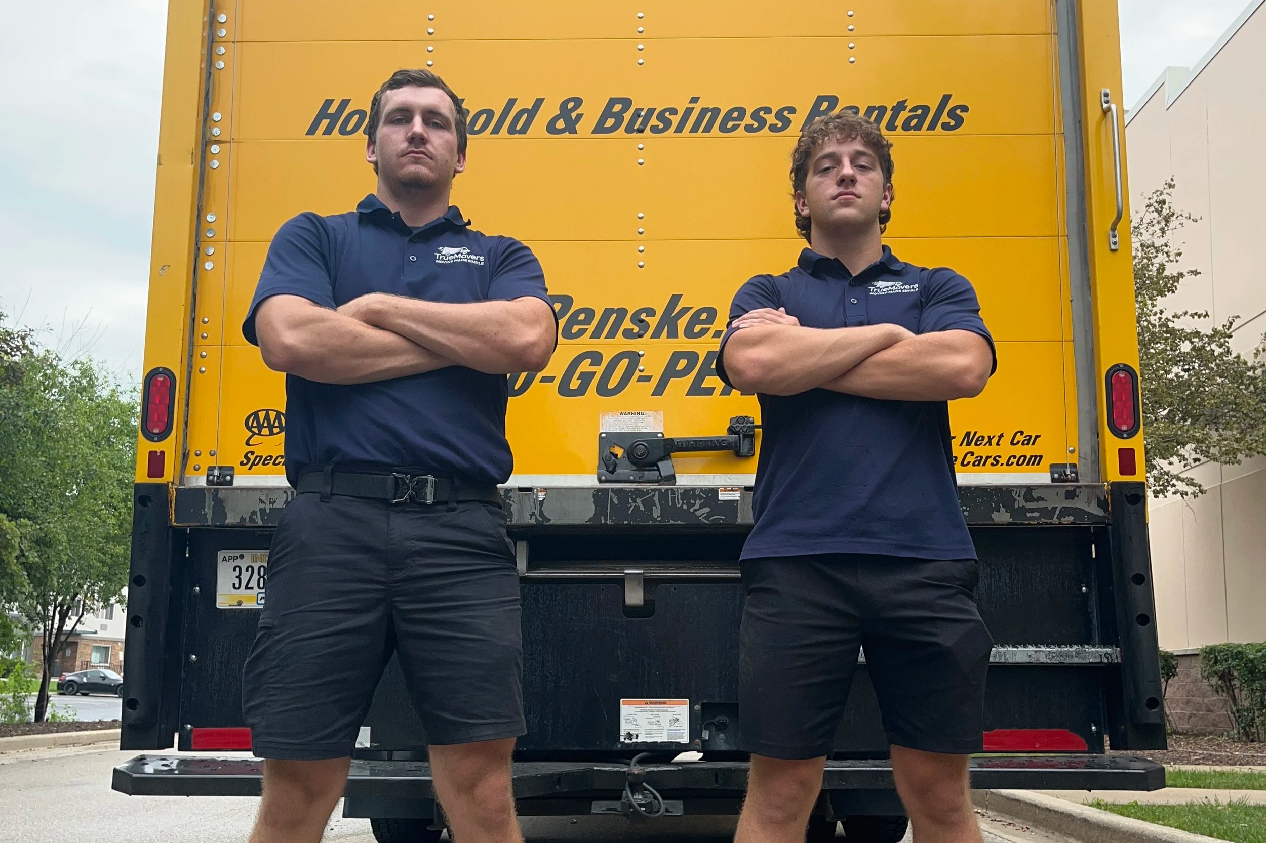Two men standing with arms crossed in front of a yellow moving truck. Both are wearing dark blue polo shirts with logos. The background shows a cloudy sky, some trees, and parked cars.