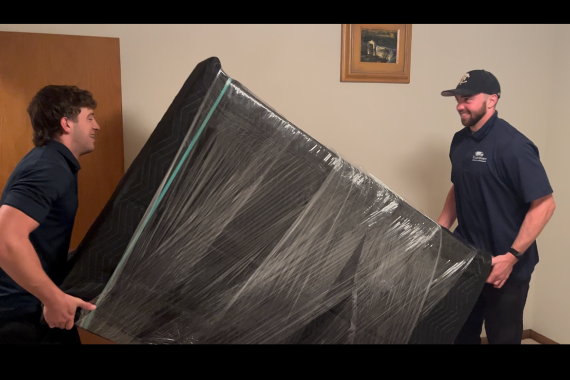 Two men are lifting and carrying a large appliance or piece of furniture wrapped in black plastic in a room with framed pictures on the wall.