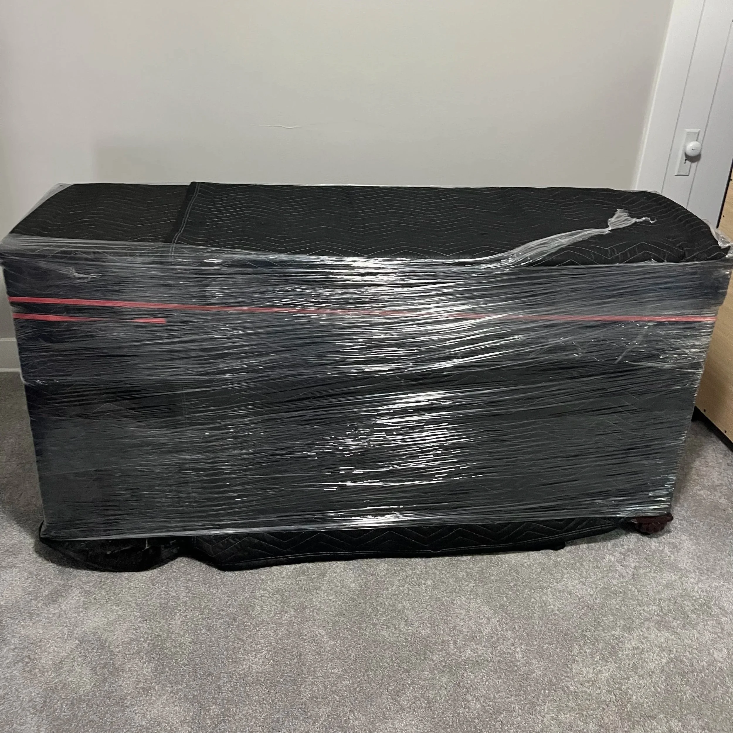 A black mattress tightly wrapped in plastic and placed on the floor in front of a white wall with a power outlet.