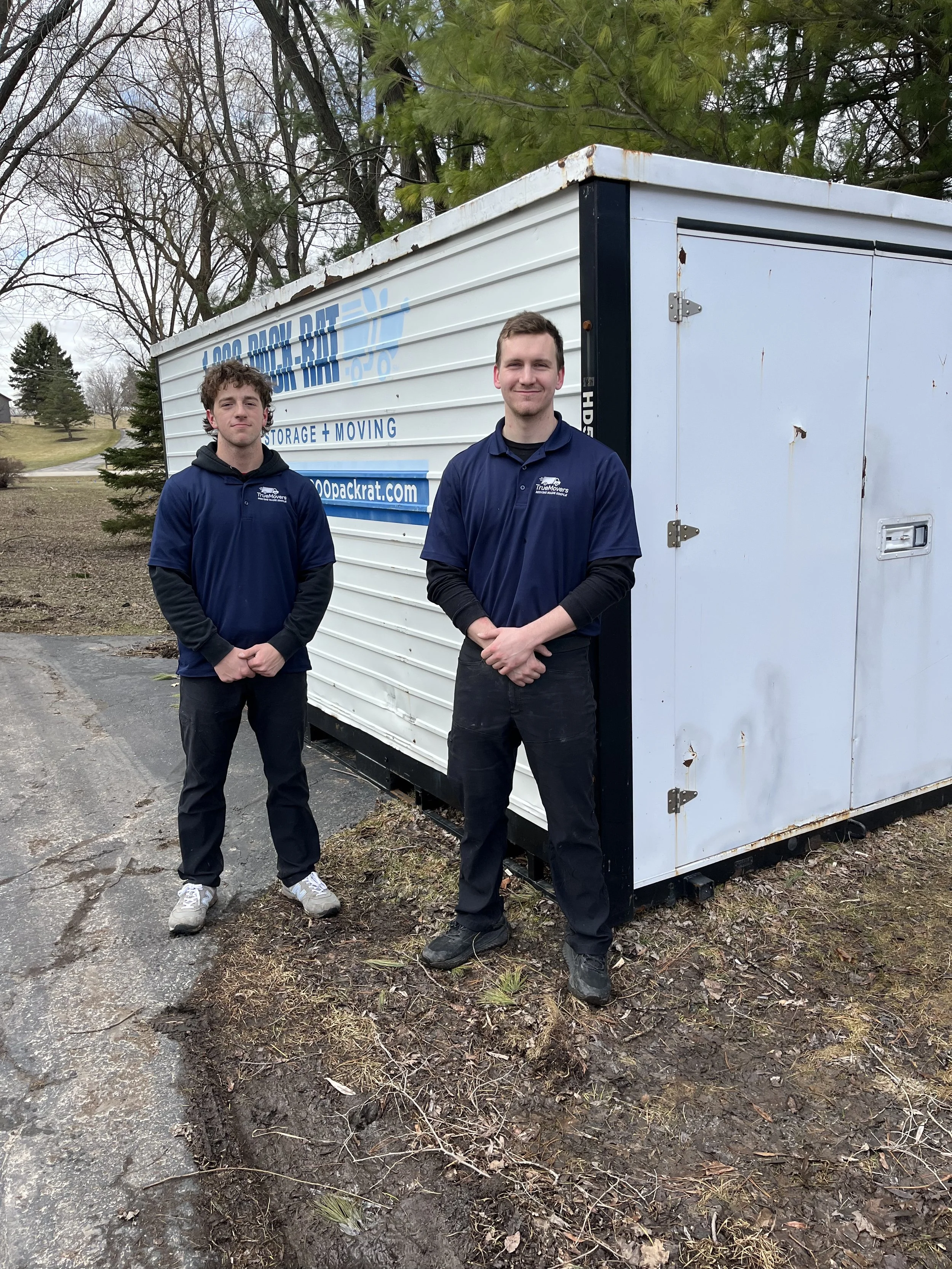 Two men standing outdoors in front of a white moving truck, with a tree line and cloudy sky in the background. They are wearing matching dark blue shirts with a company logo.