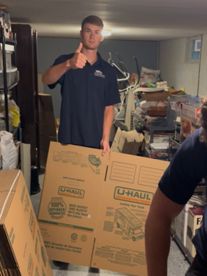 A young man stands in a cluttered basement holding a large U-Haul moving box, giving a thumbs-up.
